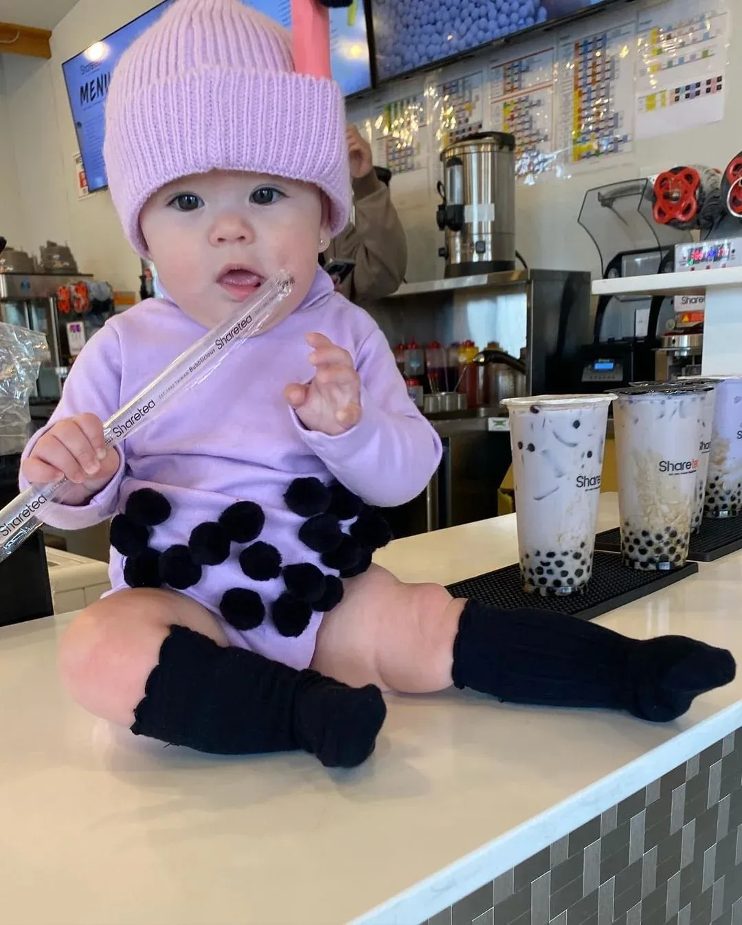 Baby in a lilac hat and outfit sitting on a counter, holding a straw at a cafe.
