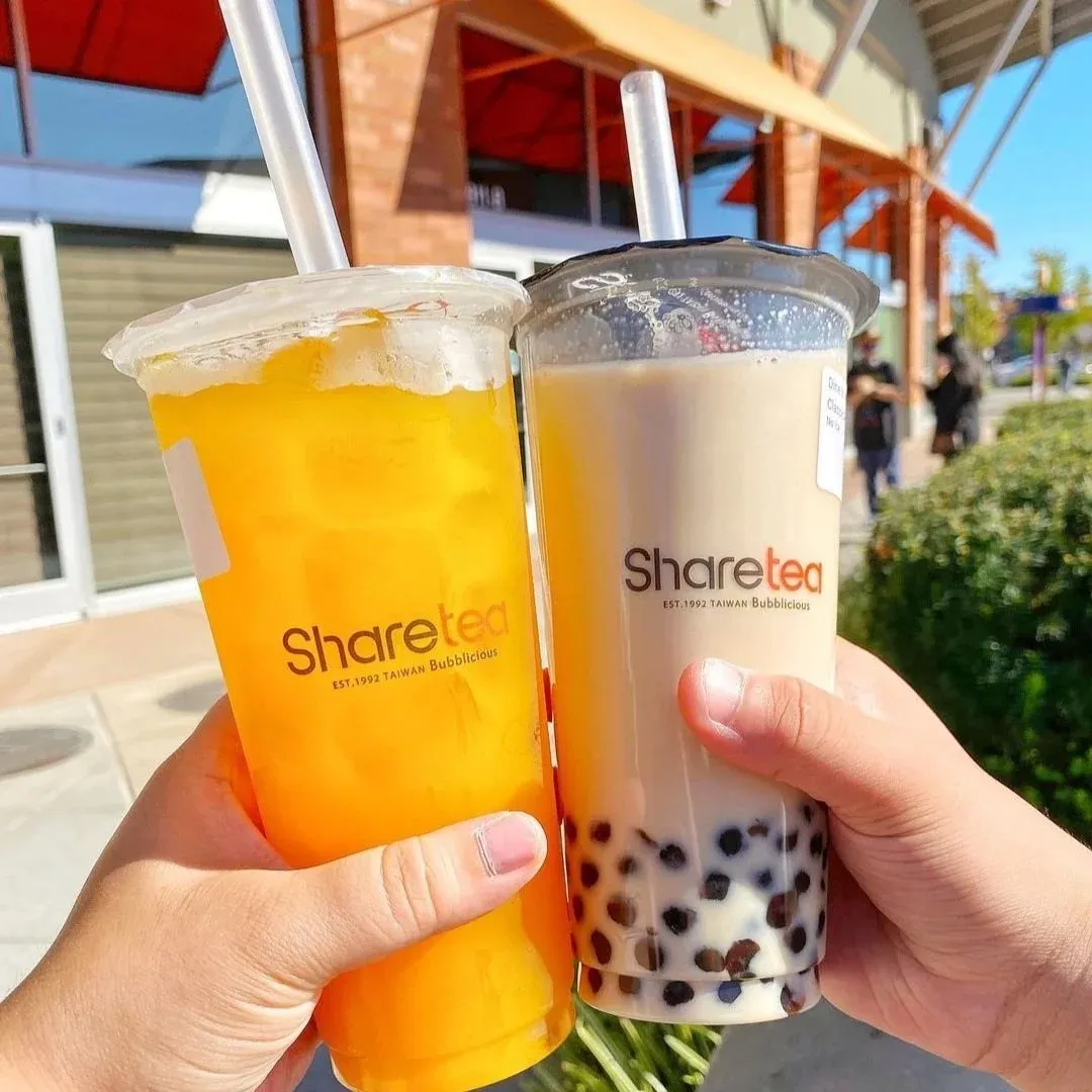 Two hands hold a mango drink and a milk tea with boba, outdoors in sunlight.