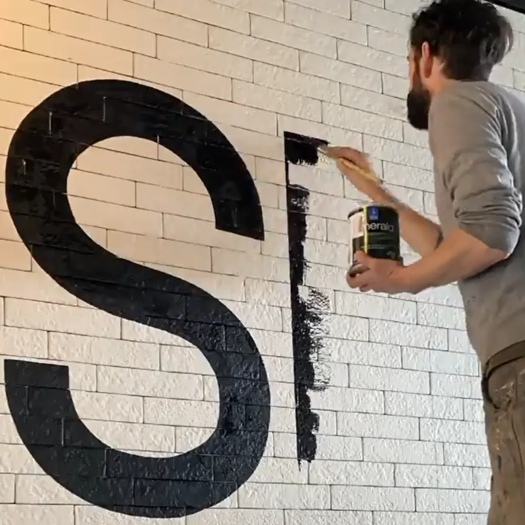 Person painting a large black letter “S” on a white brick wall using a paint can and brush.