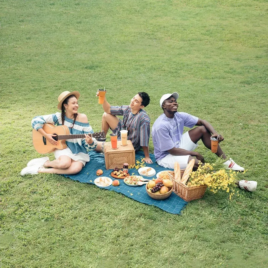 Three people picnic on a blue blanket, eating, drinking, and playing guitar on a grassy field.