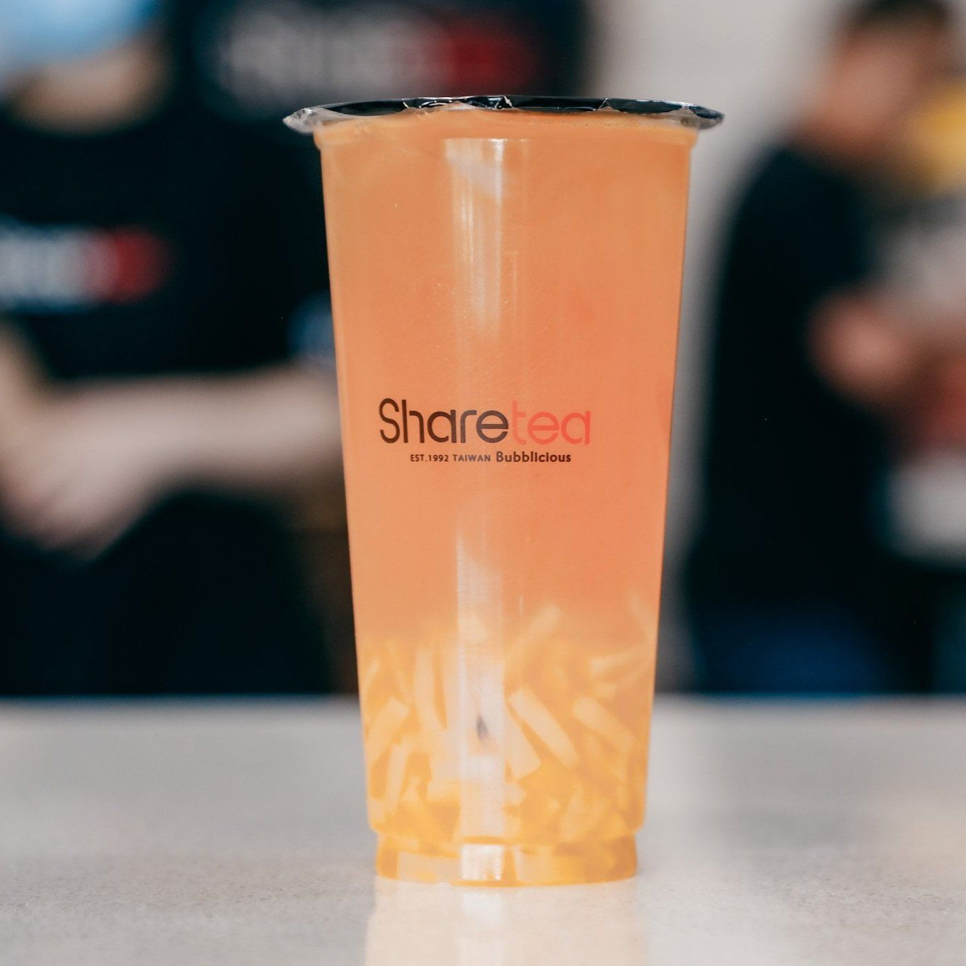 Clear plastic cup with orange drink and black lid on a table, with blurred people in the background