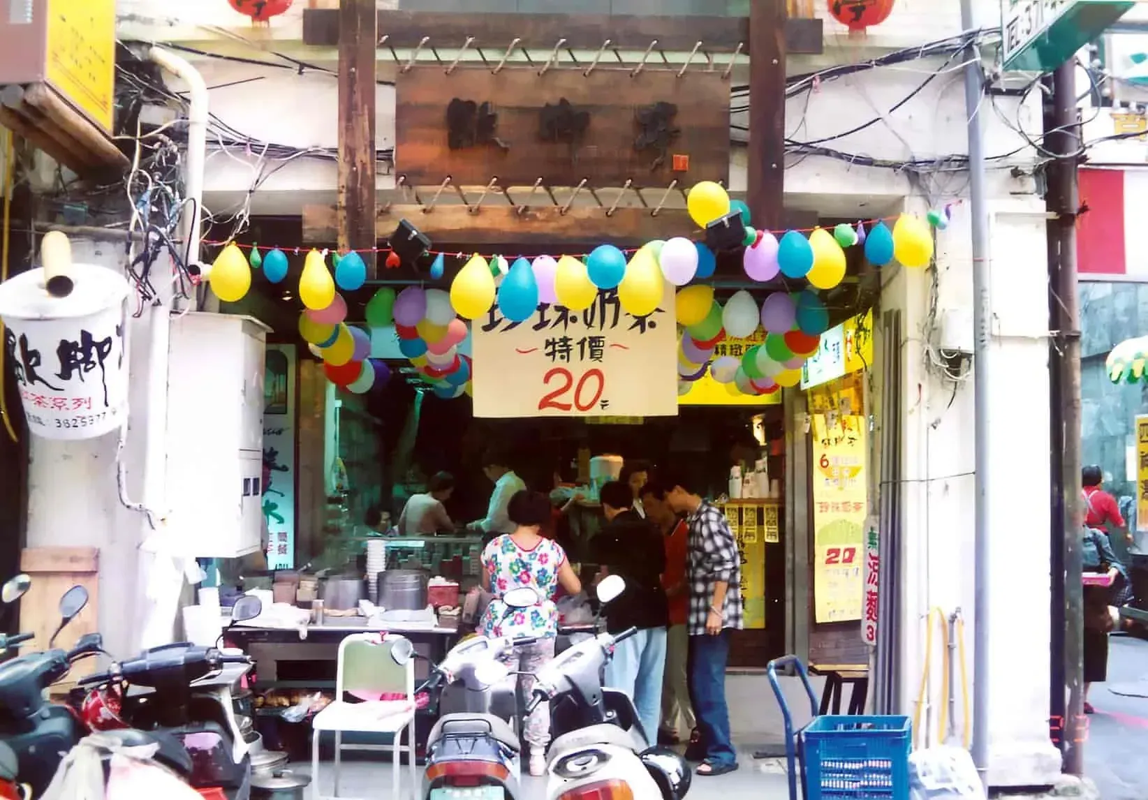 Small street shop decorated with colorful balloons, with people gathered under a sign reading “20.”