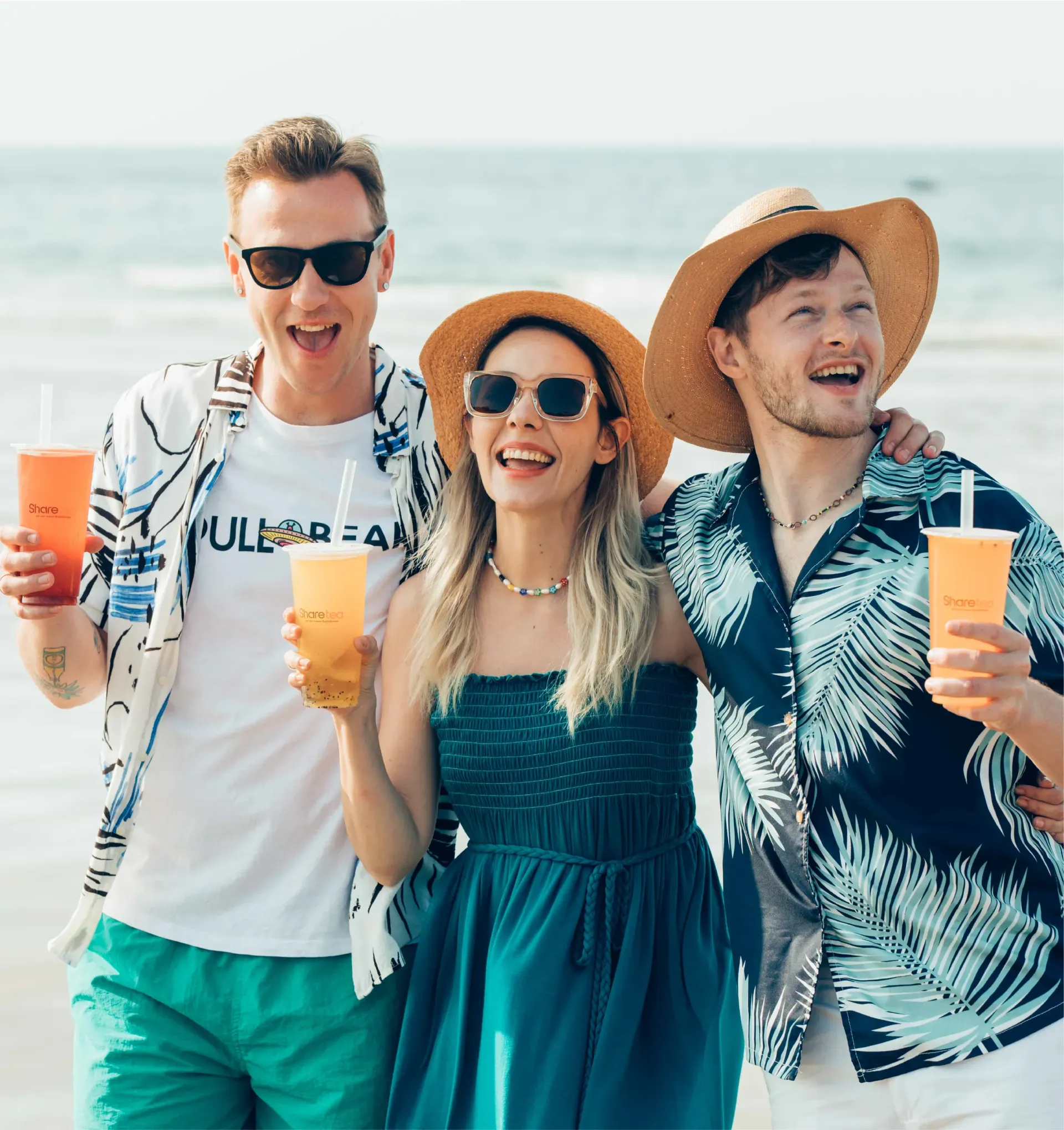 Three friends in beachwear smiling and holding drinks by the ocean