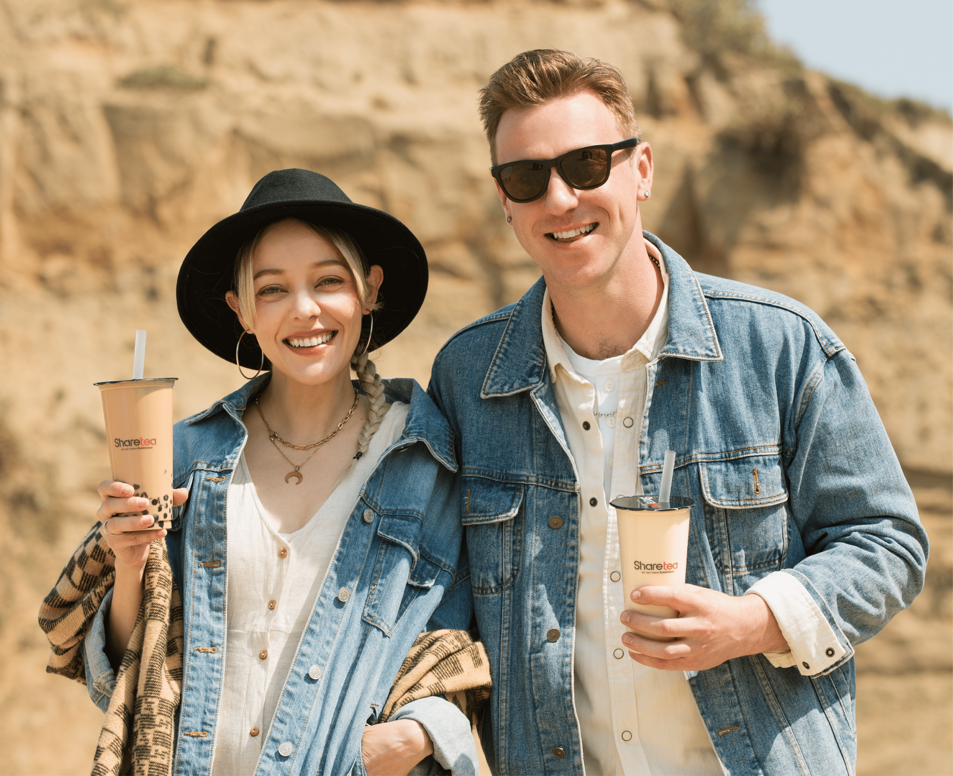 Two smiling people in denim jackets holding takeaway drinks in a rocky desert setting