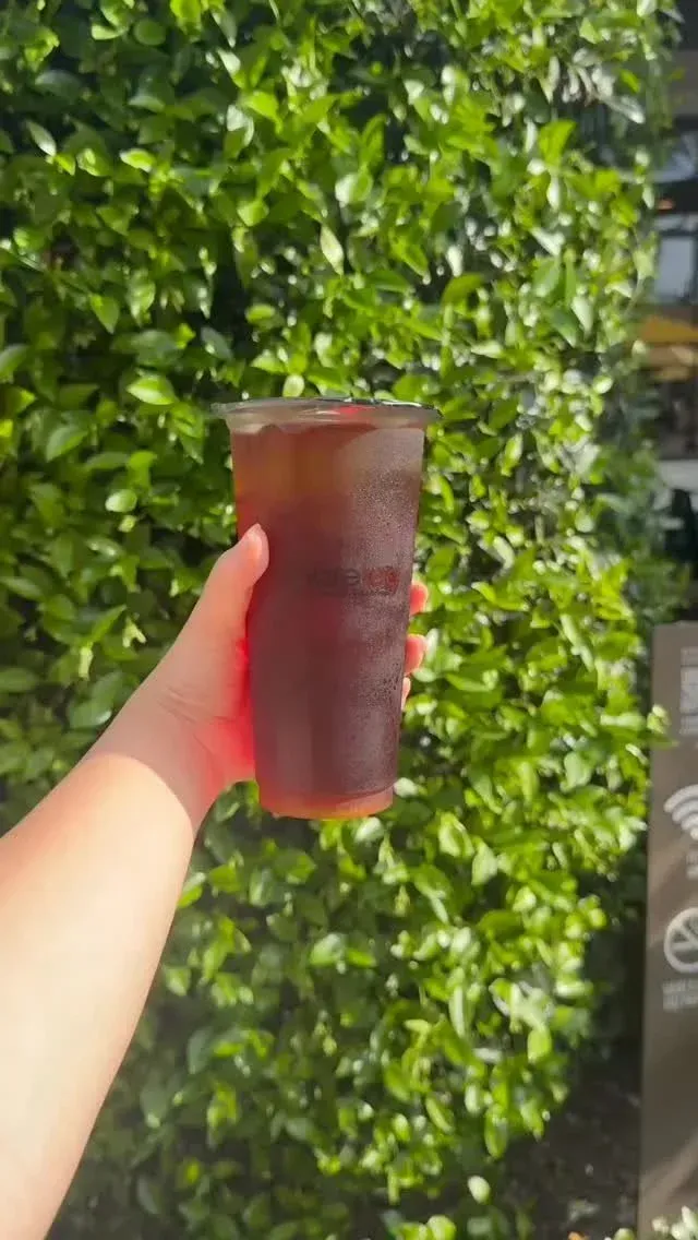 Hand holding a clear cup of iced red drink in front of green leafy plants.