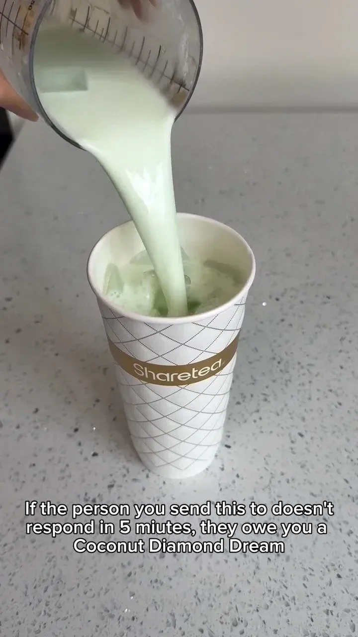 Milky green drink being poured from a measuring cup into a white patterned disposable cup