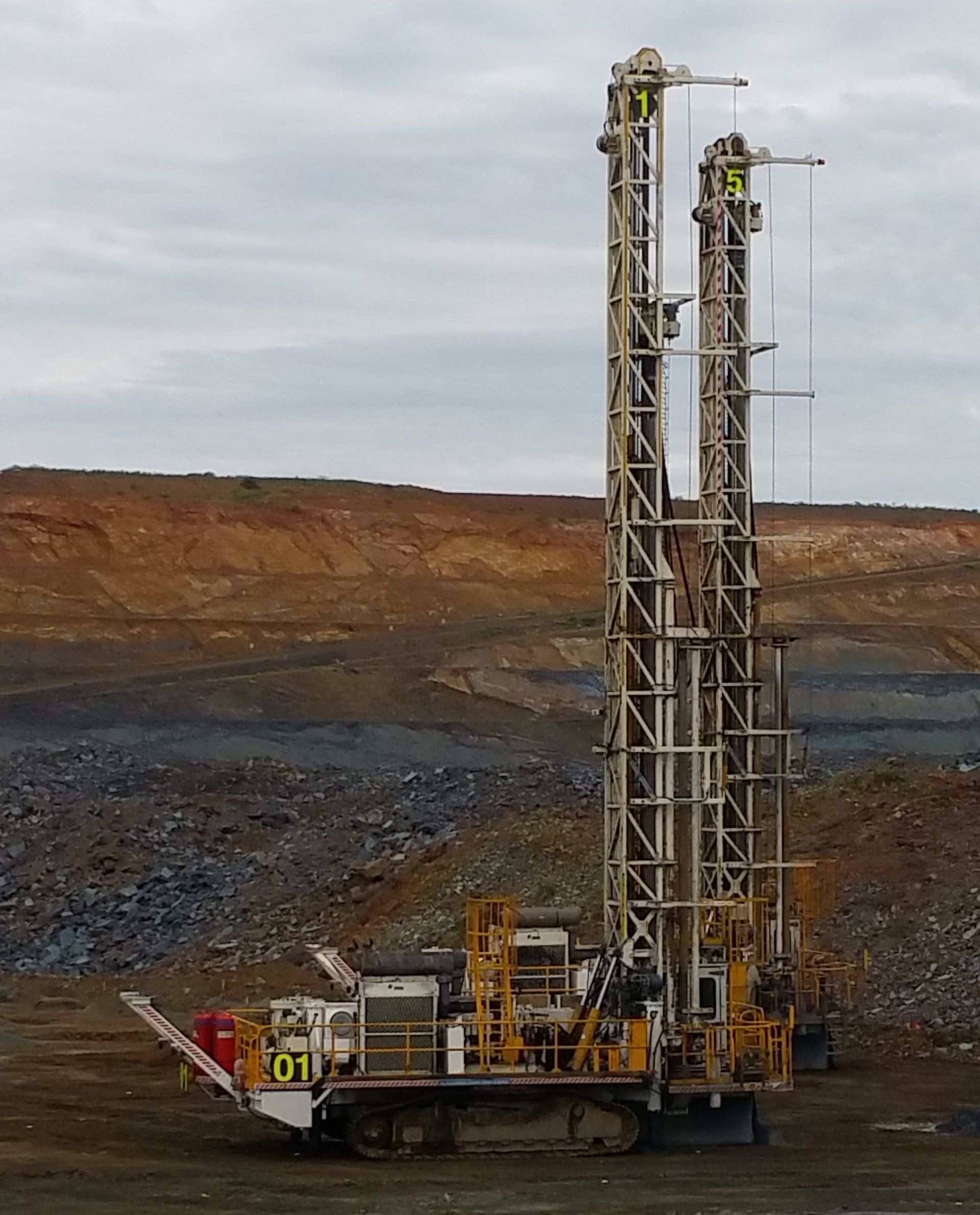 A large, yellow industrial drilling rig sits on the dirt floor of a wide, open-pit mine under a cloudy sky.