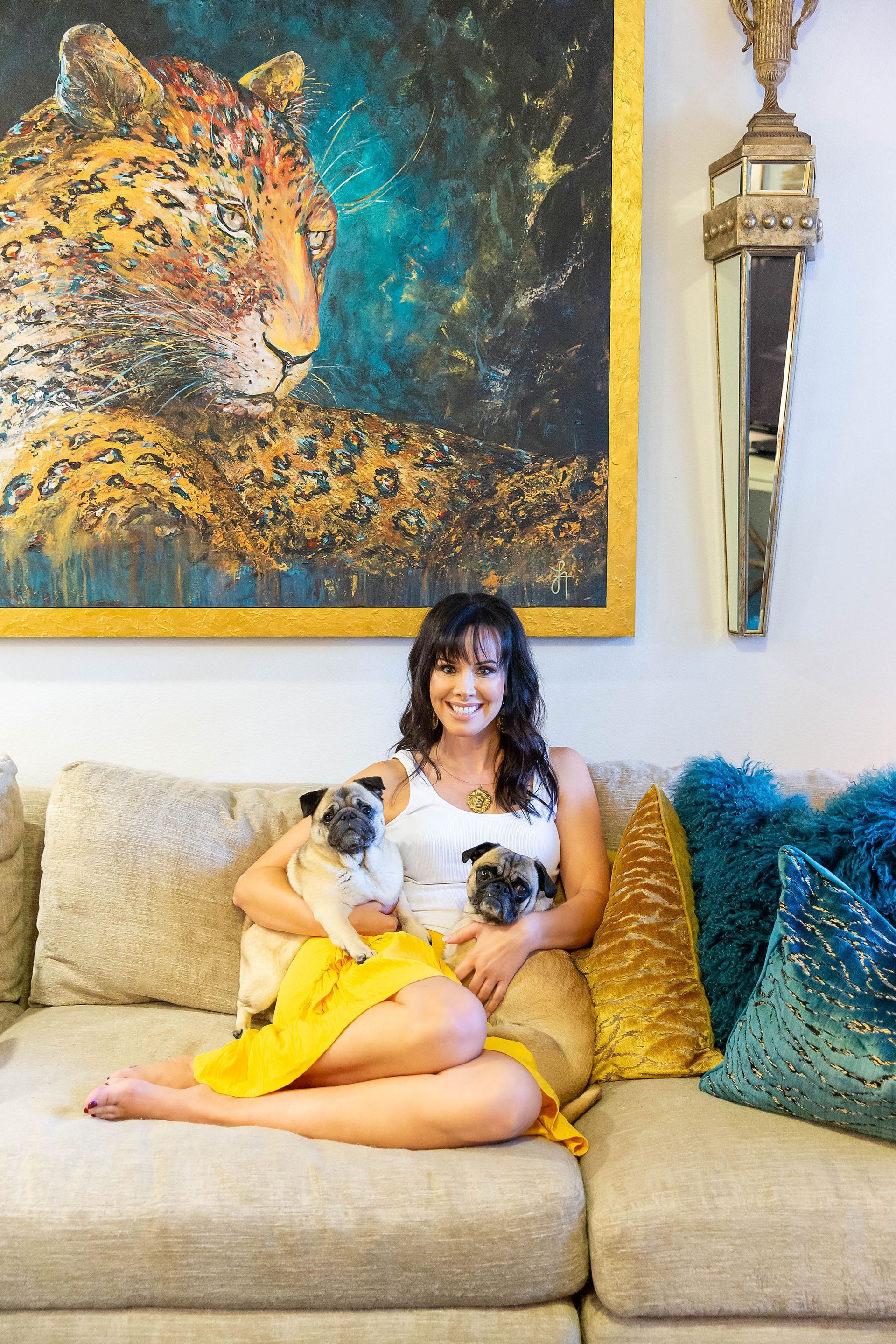 Carrie Riddle Interior Design sits on a couch with two pugs in front of a painting of a leopard