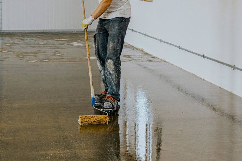 A worker in jeans and gloves applies a shiny coating to a concrete floor with a long roller.