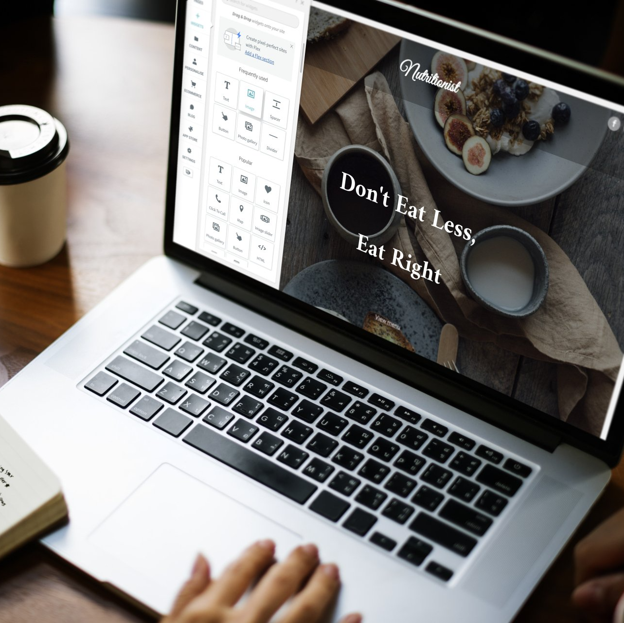 Laptop on desk with a website design being edited, coffee cup nearby, and a hand on the keyboard.
