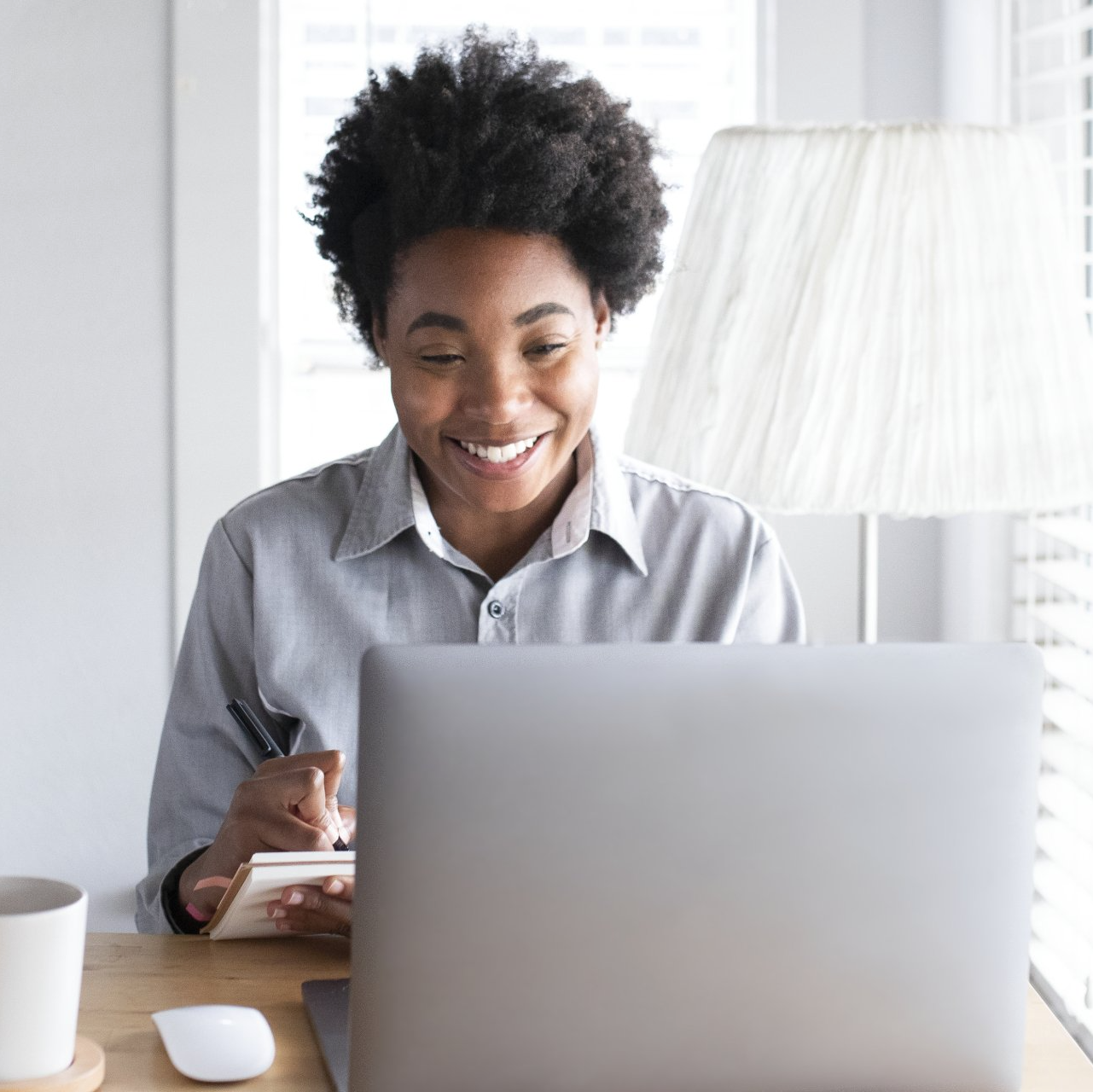 Person smiling while working on a laptop at a desk, with a notebook and lamp nearby.