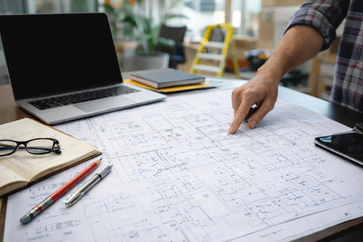 Person pointing at architectural blueprints on a desk with a laptop and drafting tools nearby