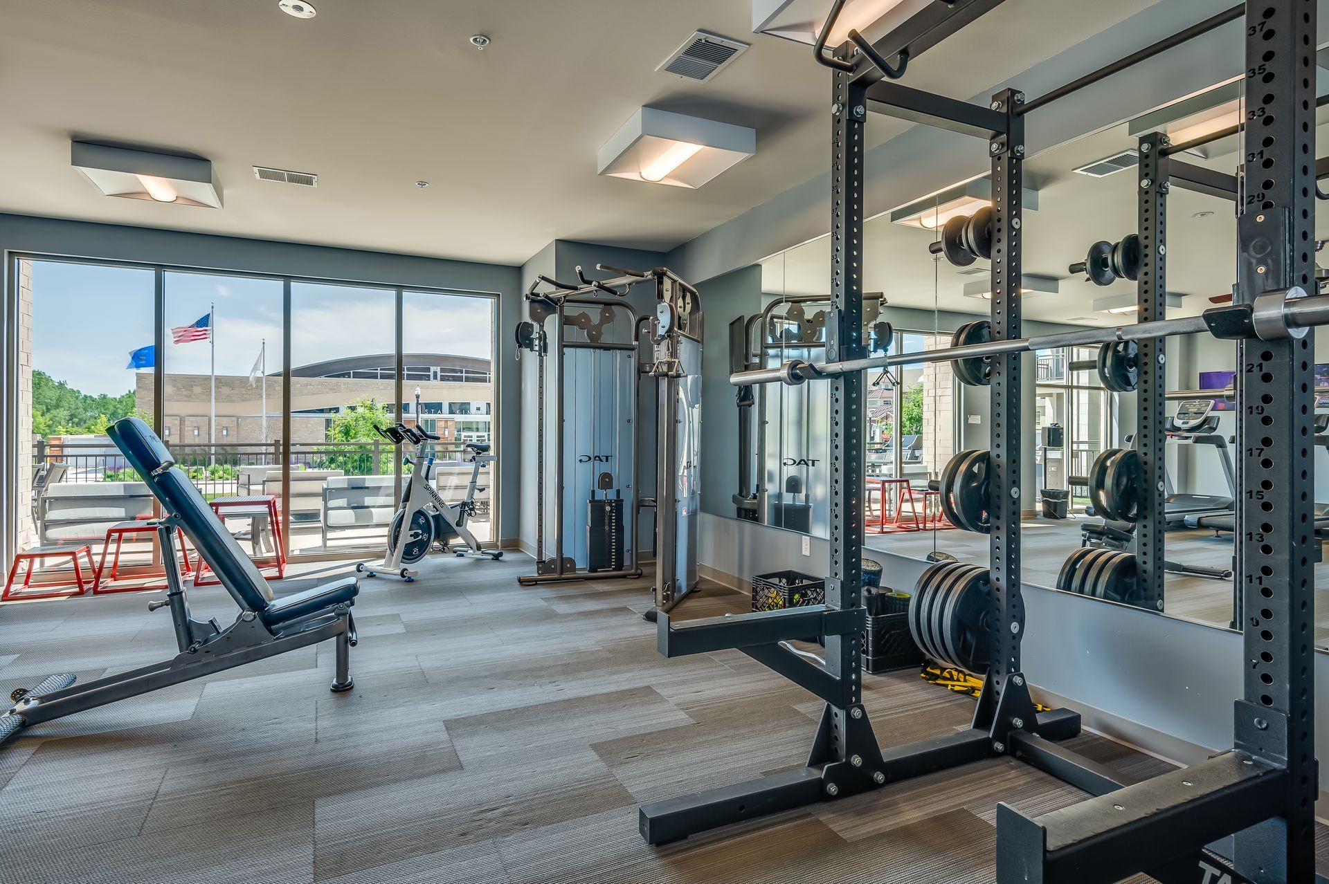 Bright gym with weight machines, mirrors, and large windows overlooking parked cars outside.