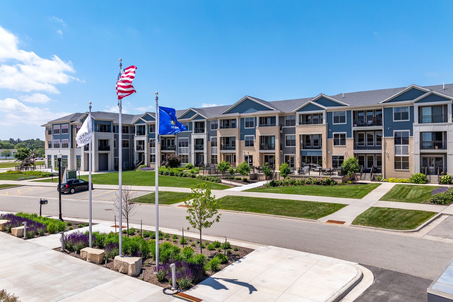 Modern apartment complex with flags, landscaped entrance, and parked cars under a blue sky