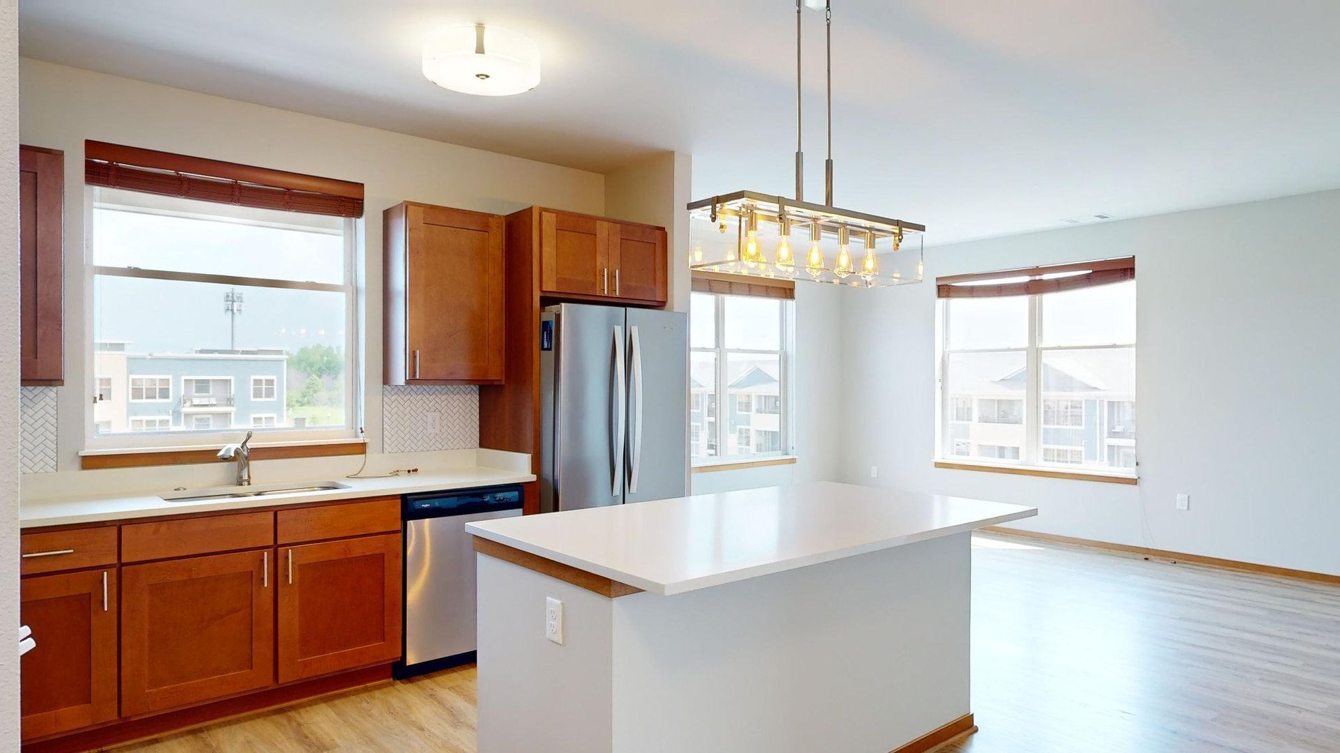Bright kitchen with wooden cabinets, stainless fridge, white island, and large windows.