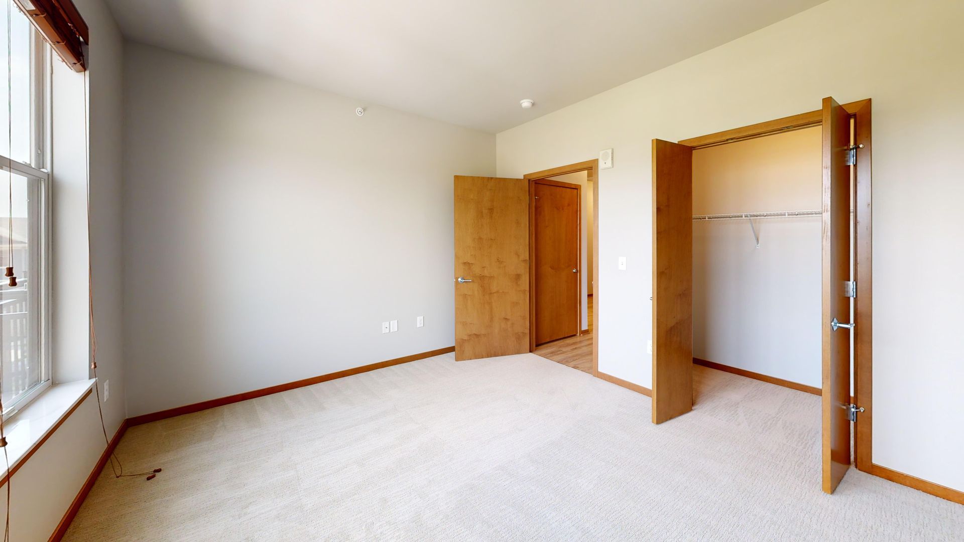 Empty beige bedroom with carpet, two open wooden closets, and a window on the left.