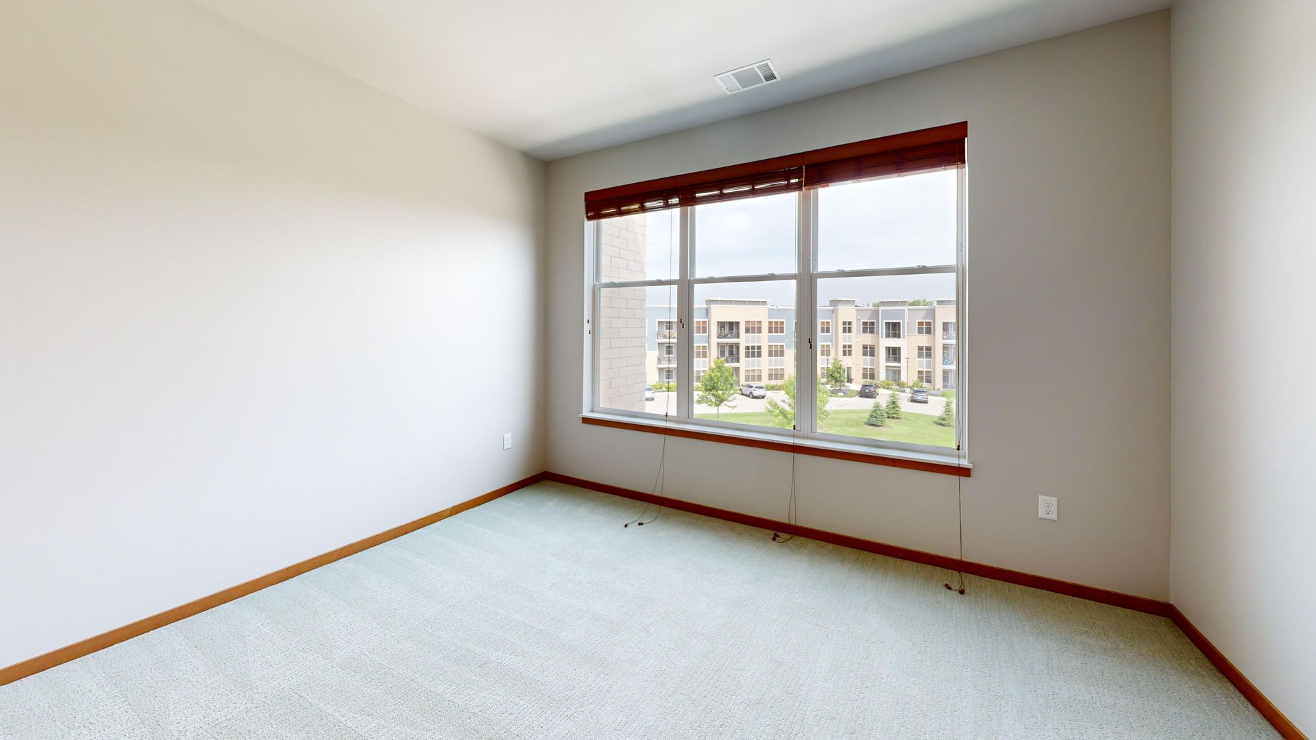 Empty bedroom with light carpet, white walls, and a large window overlooking buildings.