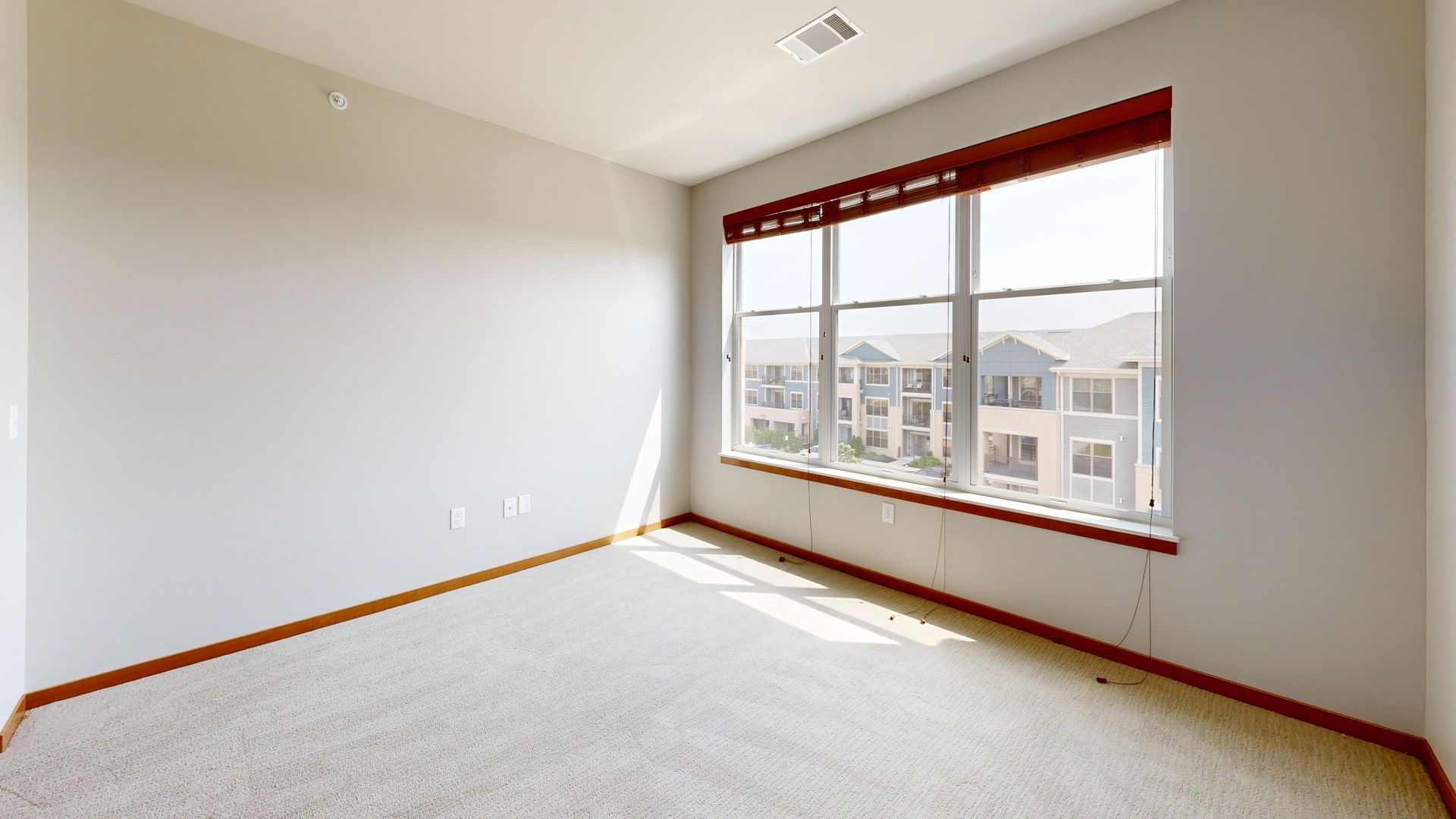 Empty sunlit bedroom with white walls, beige carpet, and a large window overlooking houses