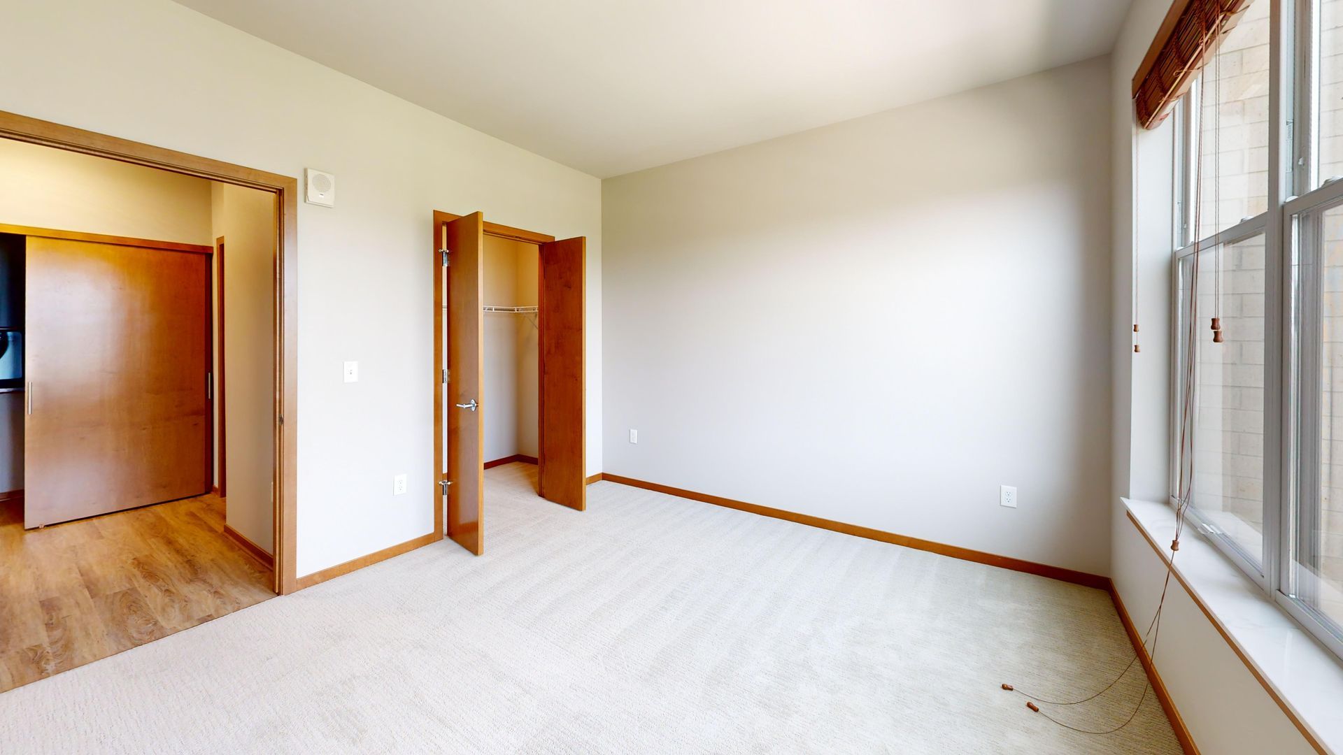 Empty beige carpeted room with white walls, wood trim, open doorway, and large windows on the right