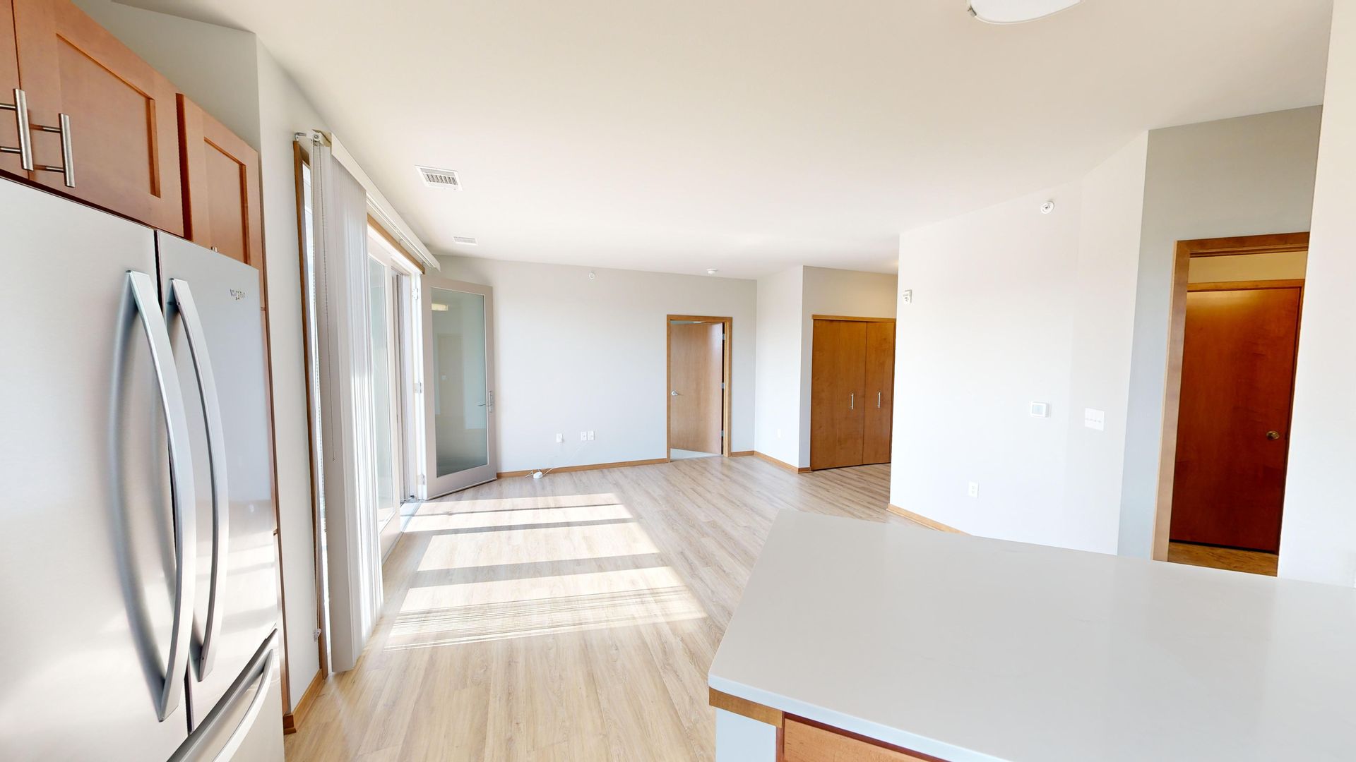 Bright empty kitchen and living area with stainless fridge, wood doors, and light wood flooring.
