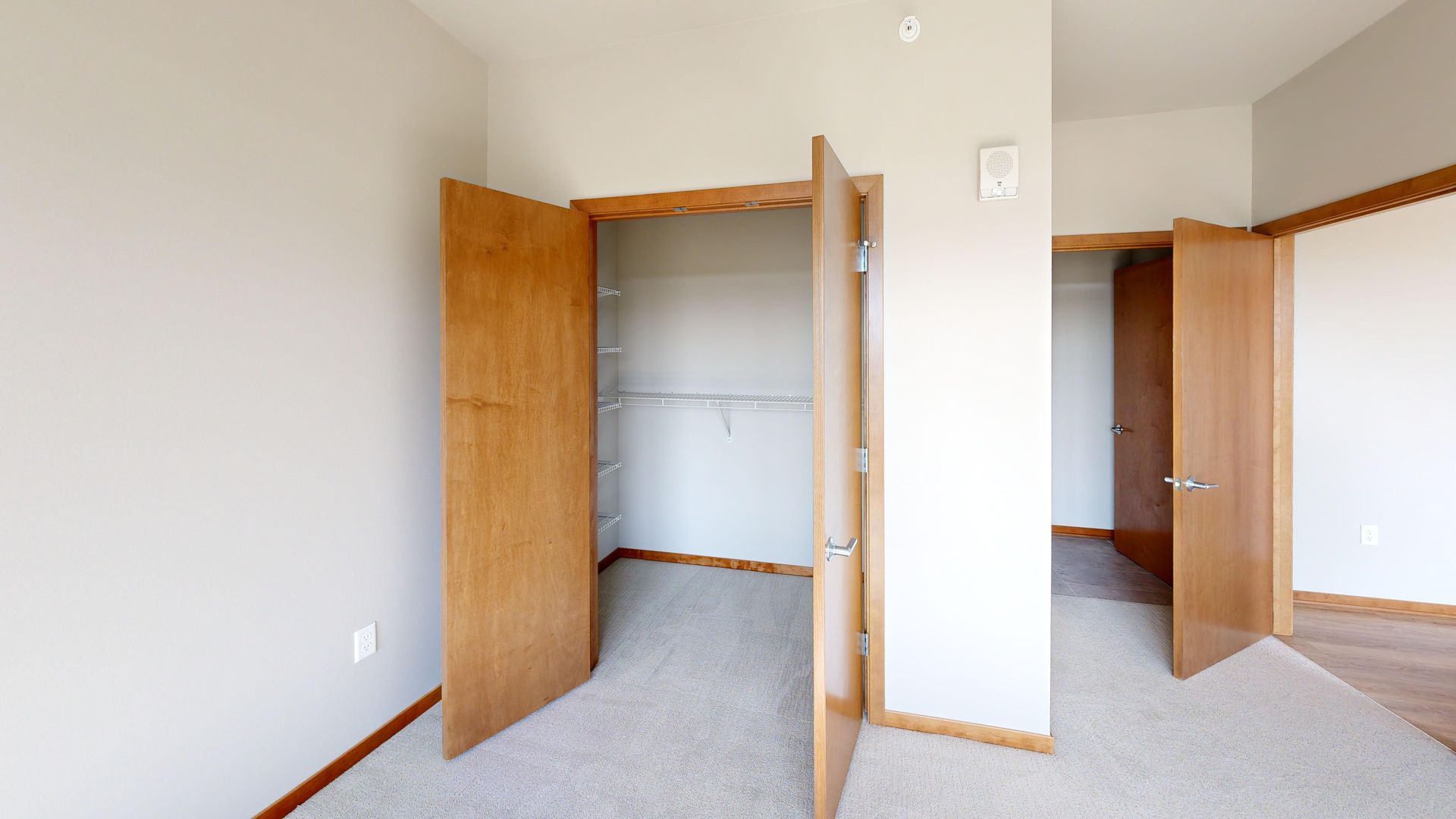 Empty beige bedroom with open wooden closet doors and gray carpet