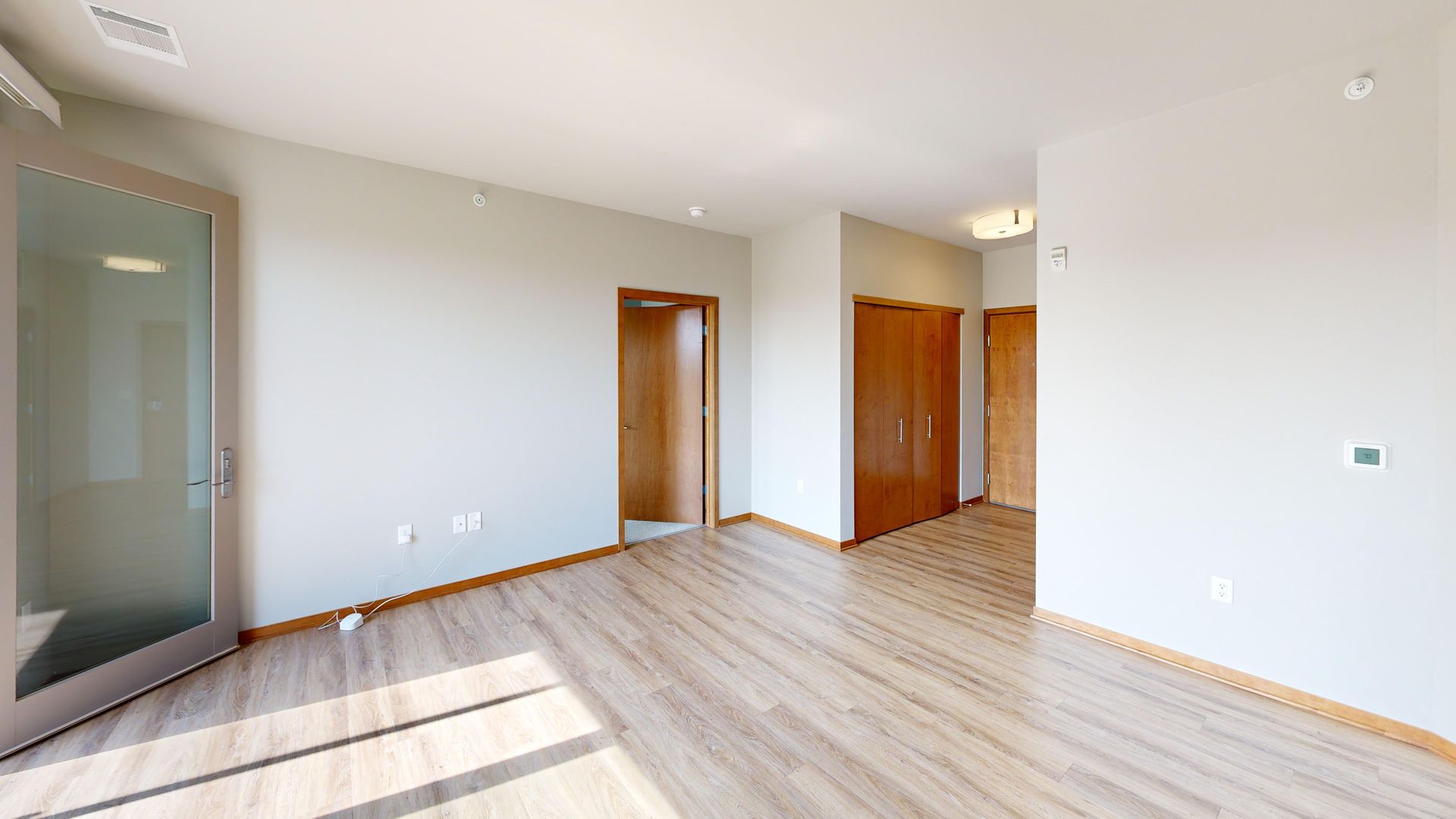 Bright empty room with light wood floors, white walls, wooden doors, and sunlight streaming in from the left.