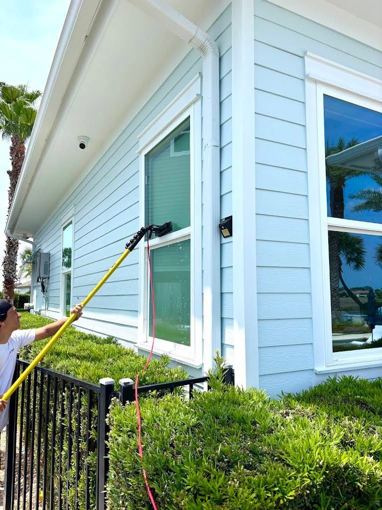Worker using a yellow pole to clean a white house’s exterior window near trimmed bushes