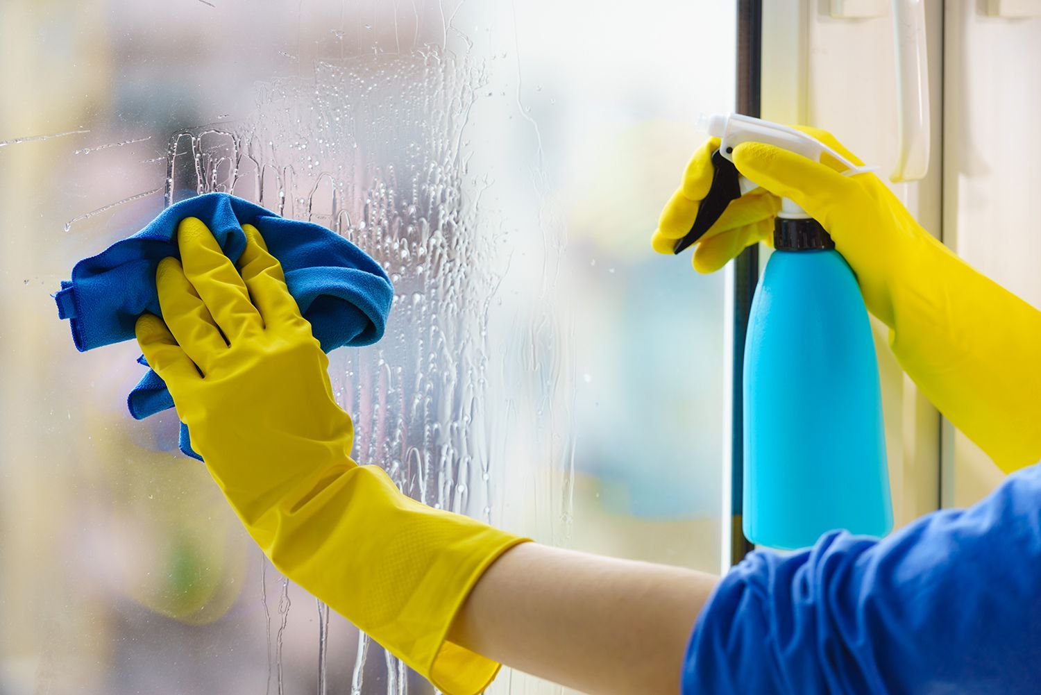 Person cleaning a window with a blue cloth and spray bottle, wearing yellow rubber gloves Person cleaning a window with a blue cloth and spray bottle, wearing yellow rubber gloves