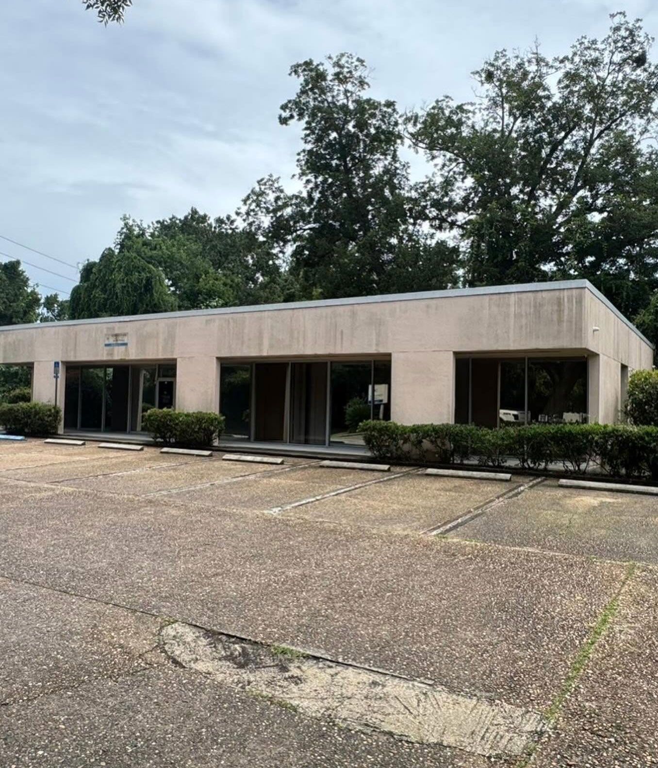 Single-story beige office building with dark windows and bushes in front, viewed from an empty parking lot