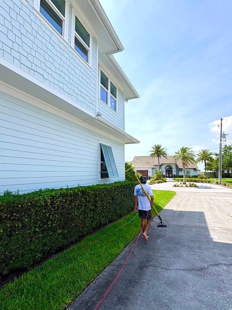 Person walking beside a white house with hedges and a sunlit driveway.