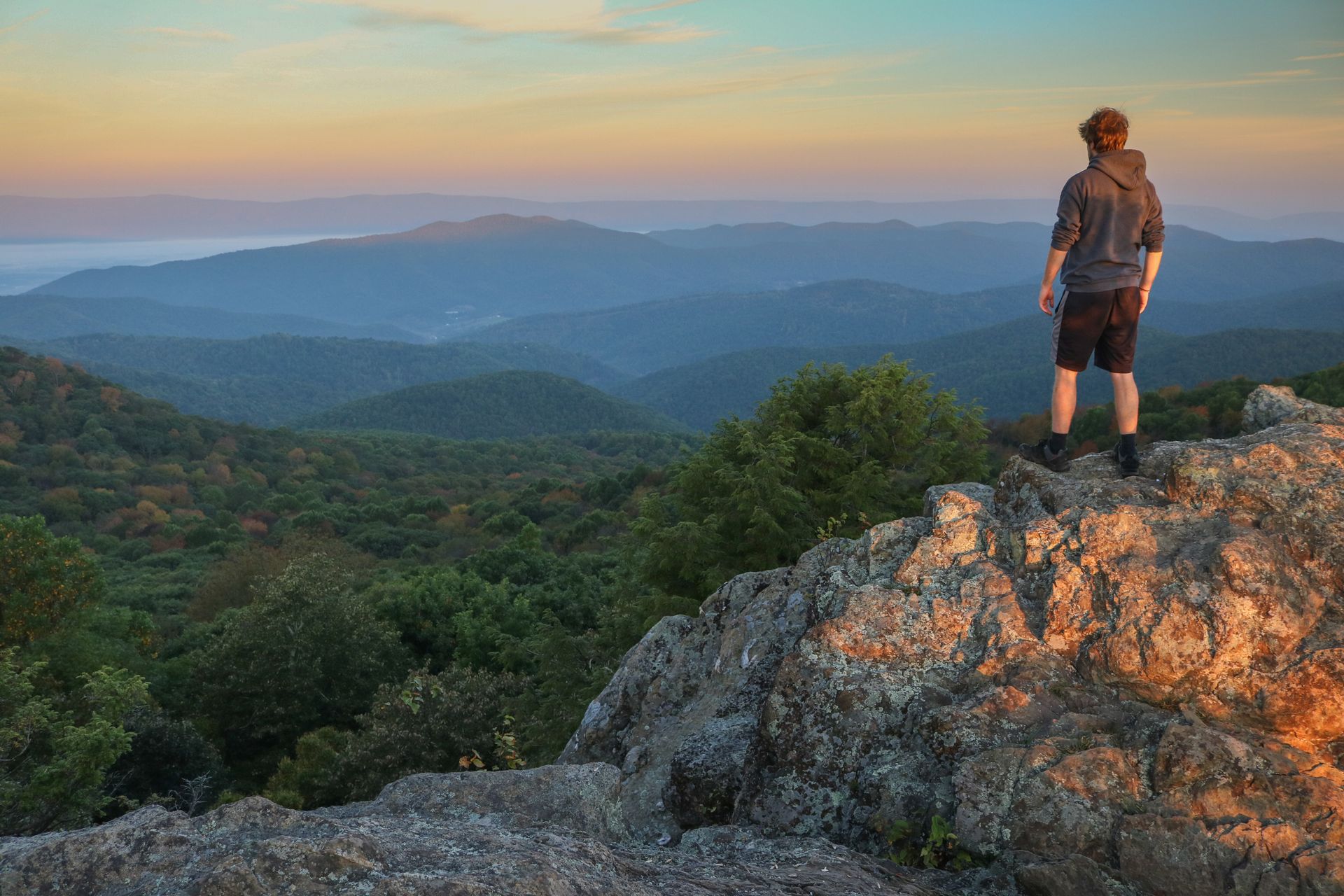 A man is standing on top of a rocky hill overlooking a mountain range.
