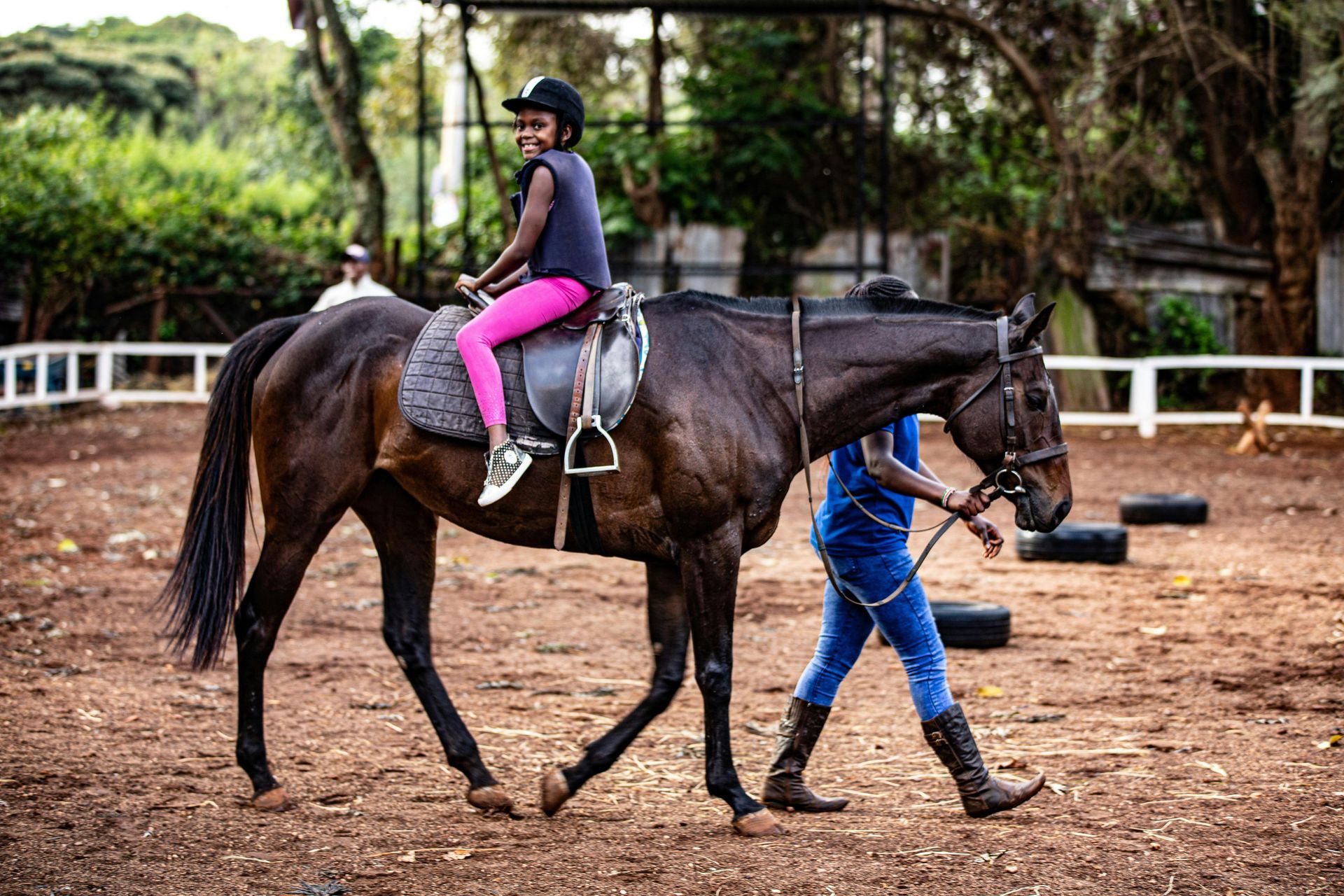 Girl riding a dark horse in an outdoor arena, led by a person in blue jeans; sunny day.