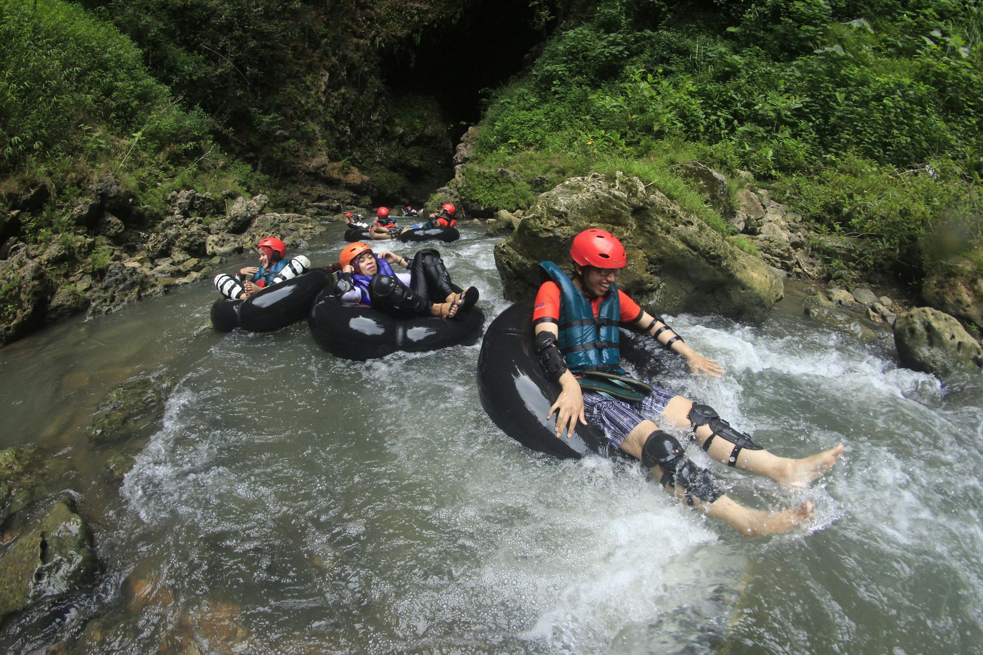People tubing down a rushing river, wearing helmets and life vests, surrounded by rocks and green foliage.