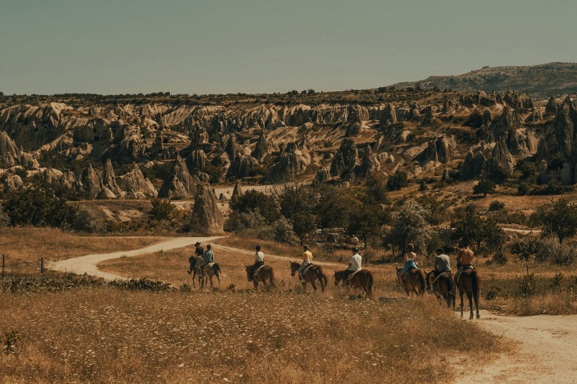 People riding horses on a dirt path through a dry, rocky landscape under a clear, sunny sky.