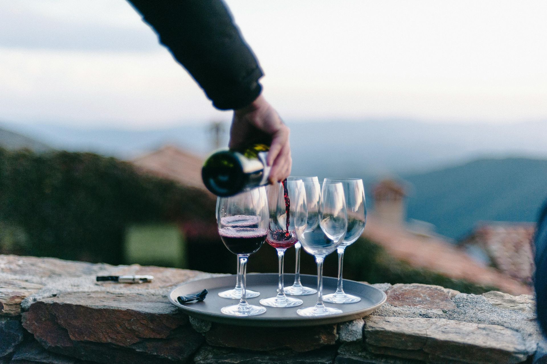 Person pouring red wine into glasses on a tray, outdoors with a mountain backdrop.