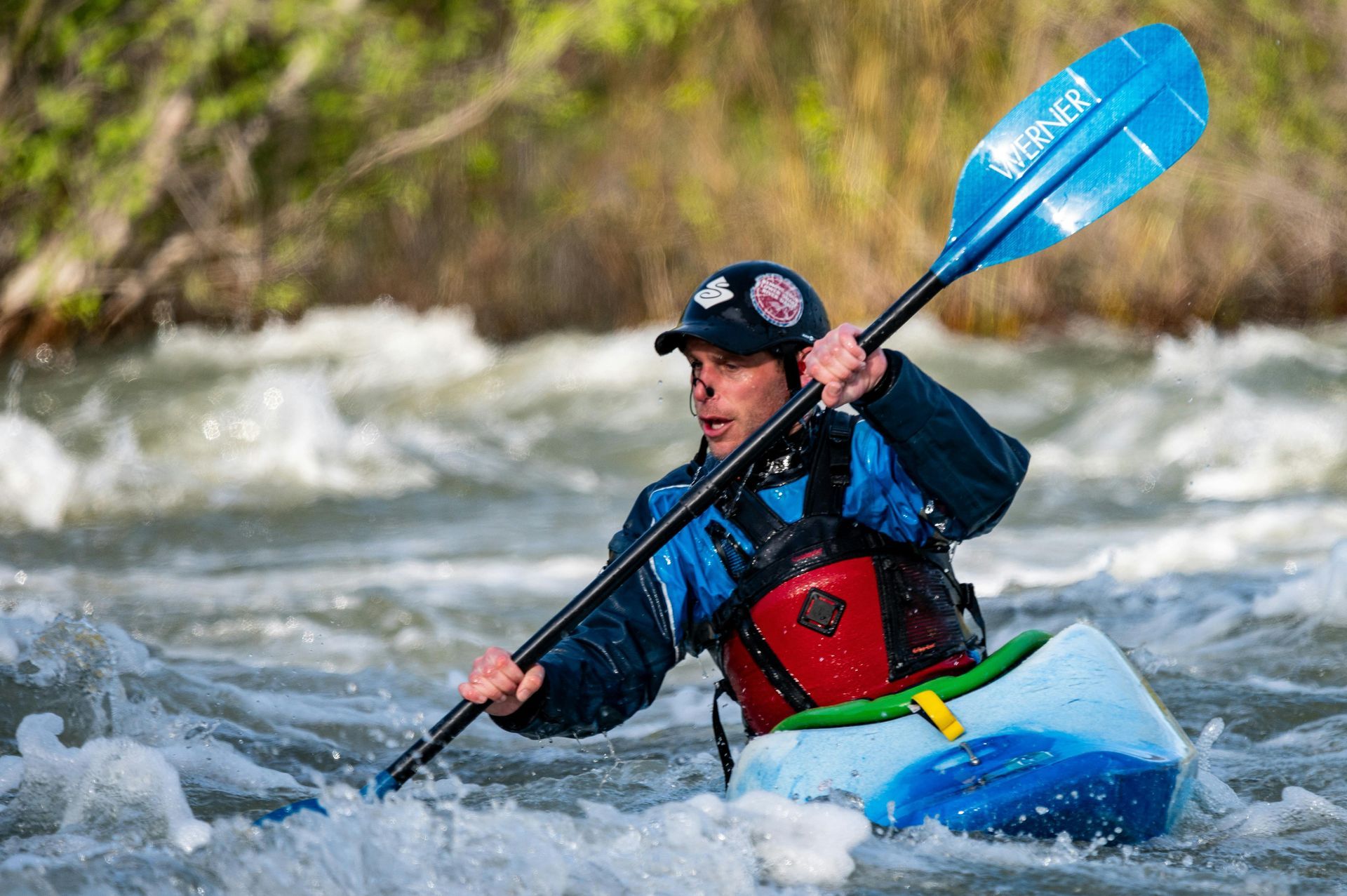 Kayaker paddles in turbulent water, wearing safety gear. Blue kayak, paddle, and sky.