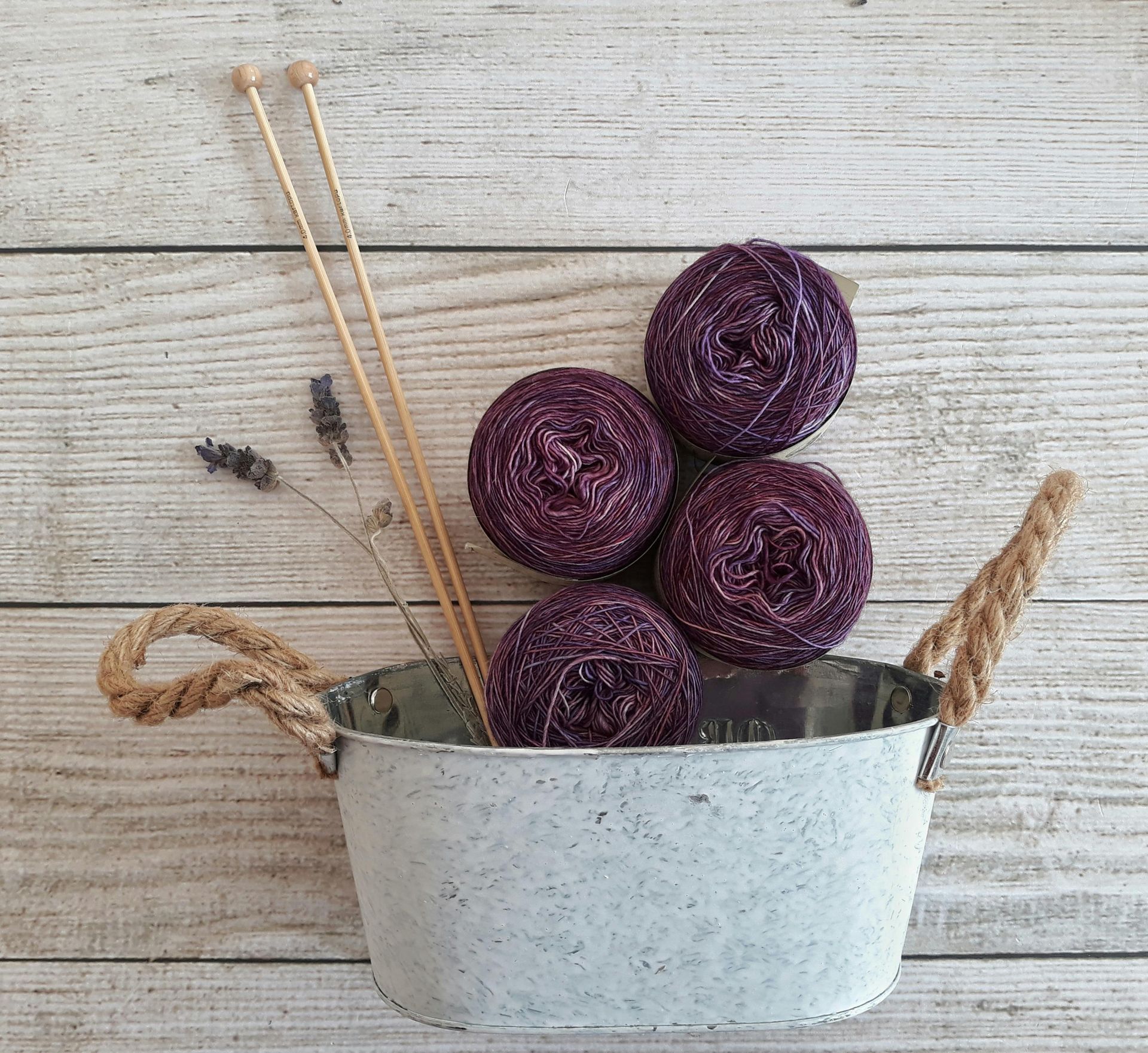 Four balls of purple yarn, needles, and dried lavender in a metal tub on a wooden surface.
