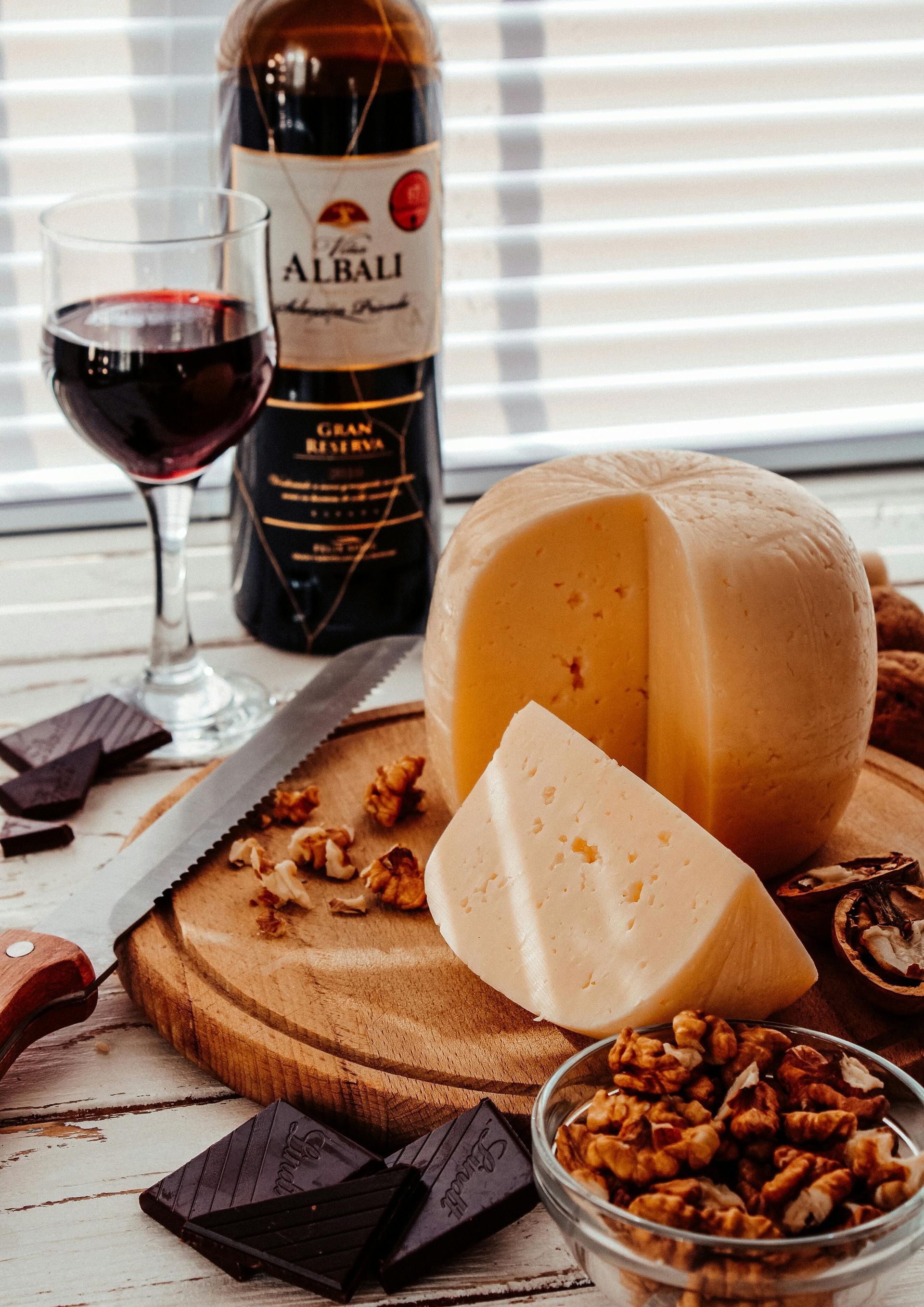 A bottle of wine , cheese , chocolate , nuts and a glass of wine are on a table.