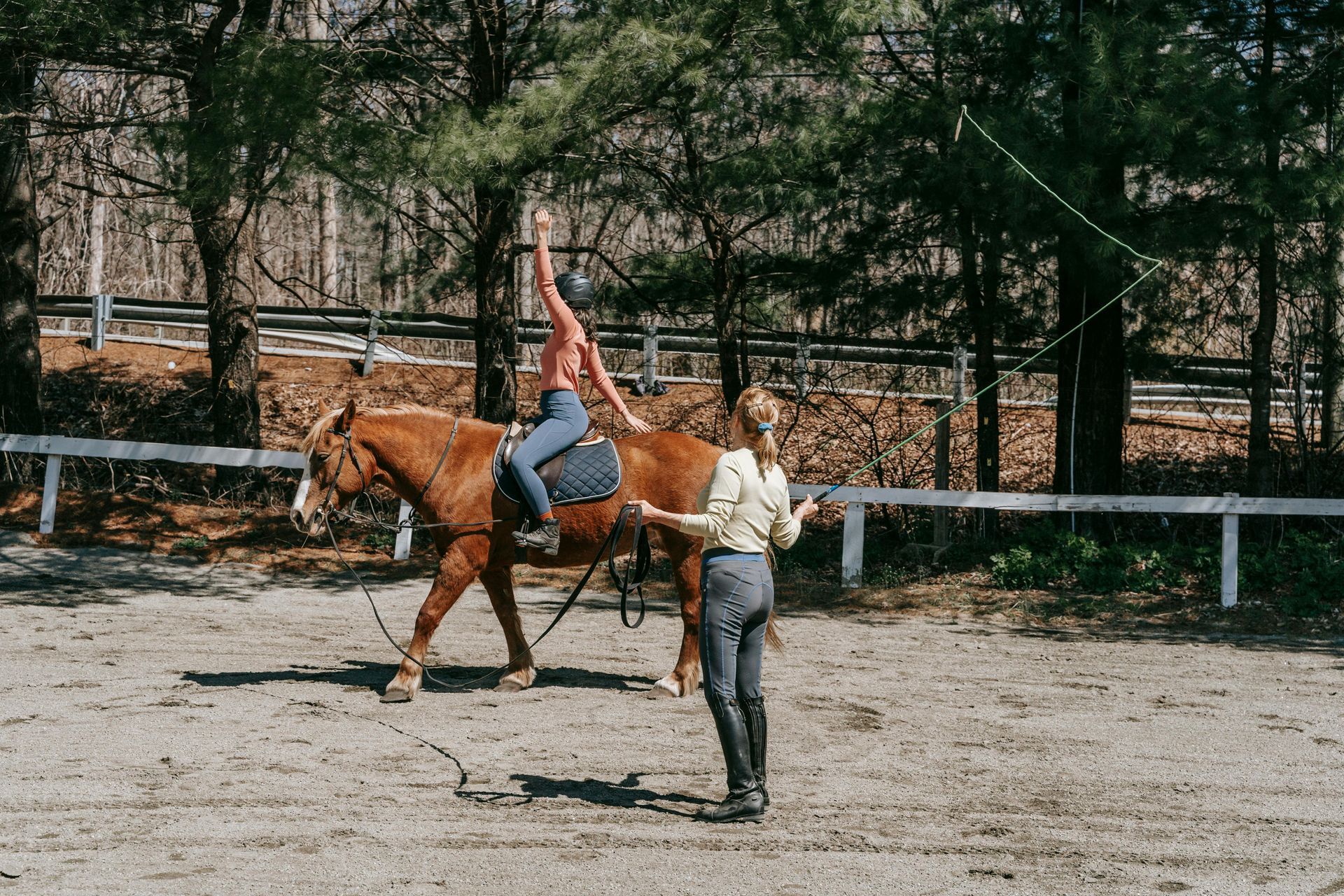 Woman on horseback, arms raised, with trainer in outdoor ring holding a lunge whip.