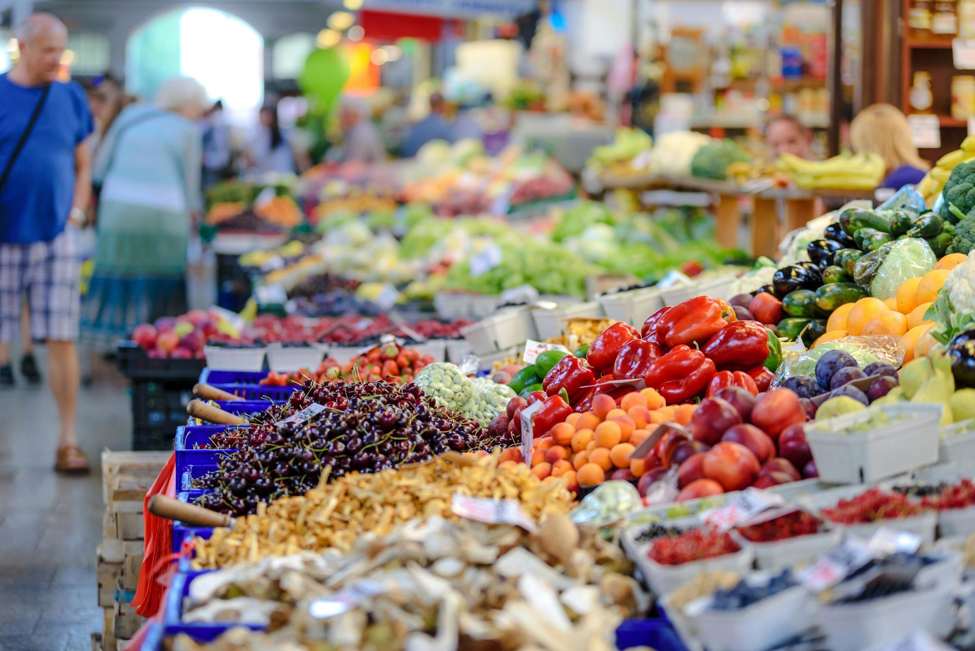 Market with colorful produce: peppers, berries, and more. People browse in a sunny, outdoor setting.