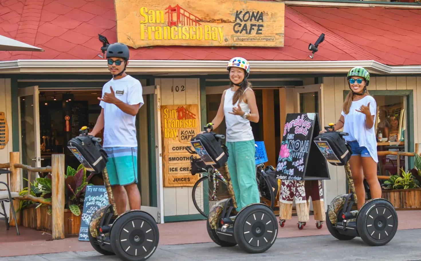 Three people ride Segways past a colorful storefront with a yellow awning and bicycles parked outside.