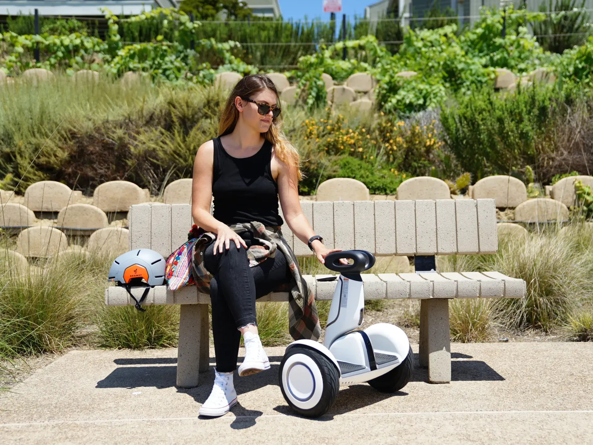 Woman in black seated on a park bench beside a white scooter, with greenery and stone landscaping behind her
