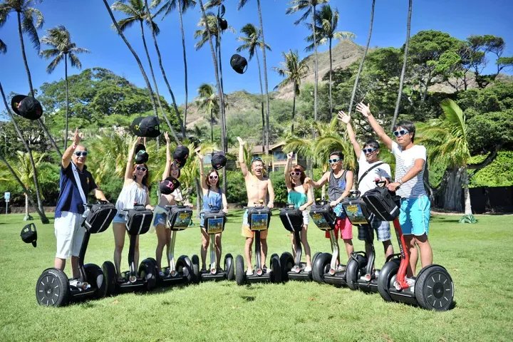 Group riding Segways on a sunny tropical lawn with palm trees, smiling and raising their arms