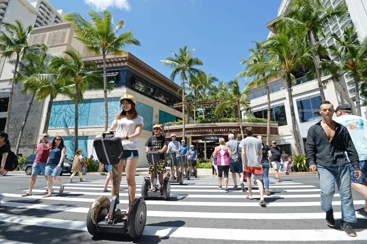 People riding scooters and pedestrians crossing a sunny striped crosswalk in a palm-lined shopping area