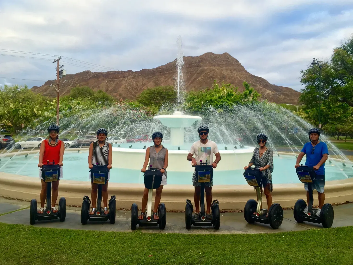 Six people standing on Segways in front of a fountain with mountains and trees behind them.