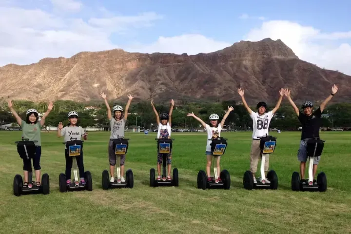 Seven people on Segways with arms raised, standing on grass in front of mountains.