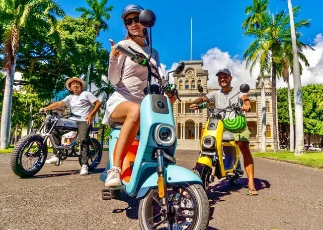 People riding colorful e-scooters on a sunny street with palm trees and a historic building in the background