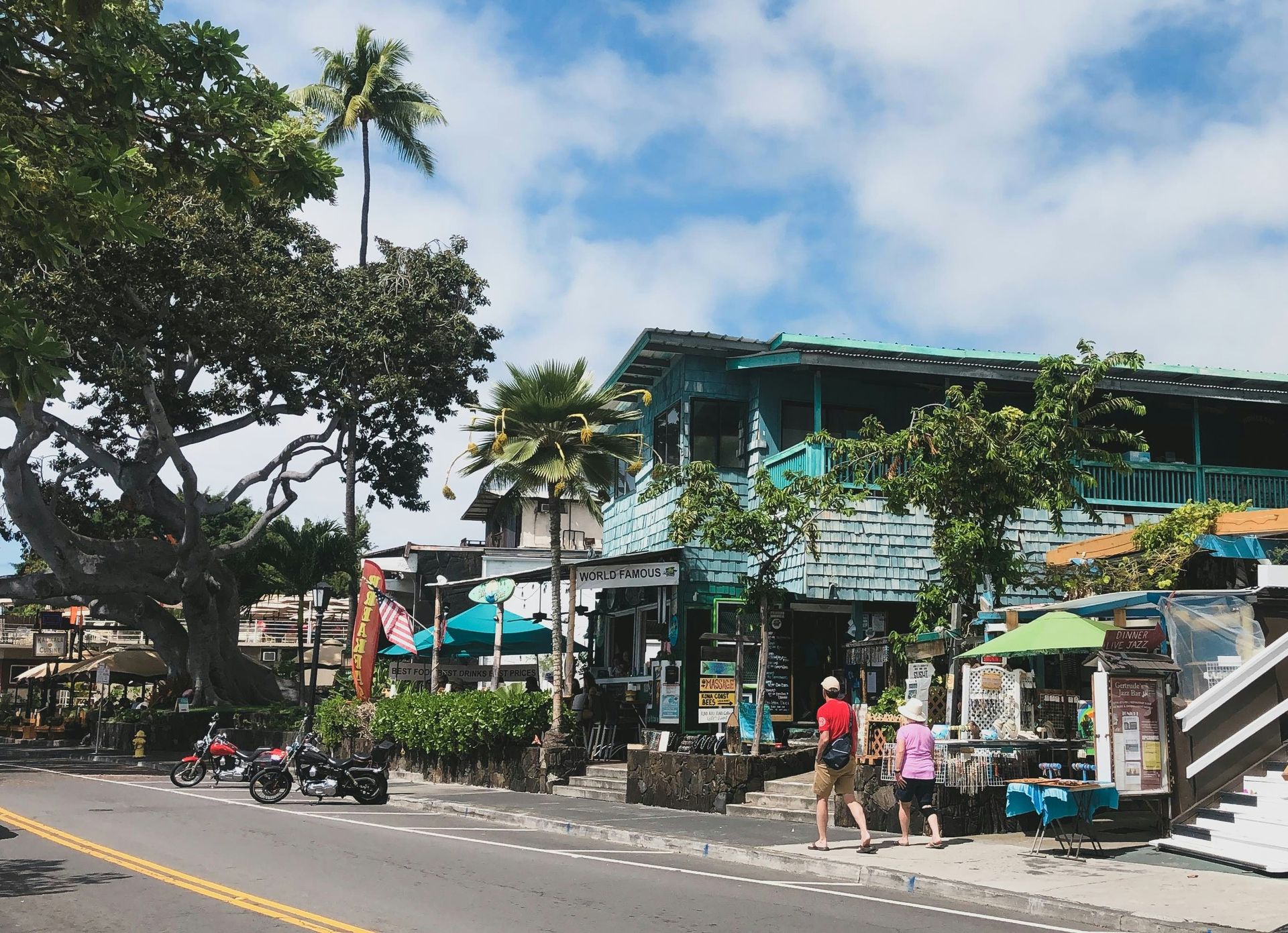 Street market with shops, umbrellas, pedestrians, and a large tree under a partly cloudy sky