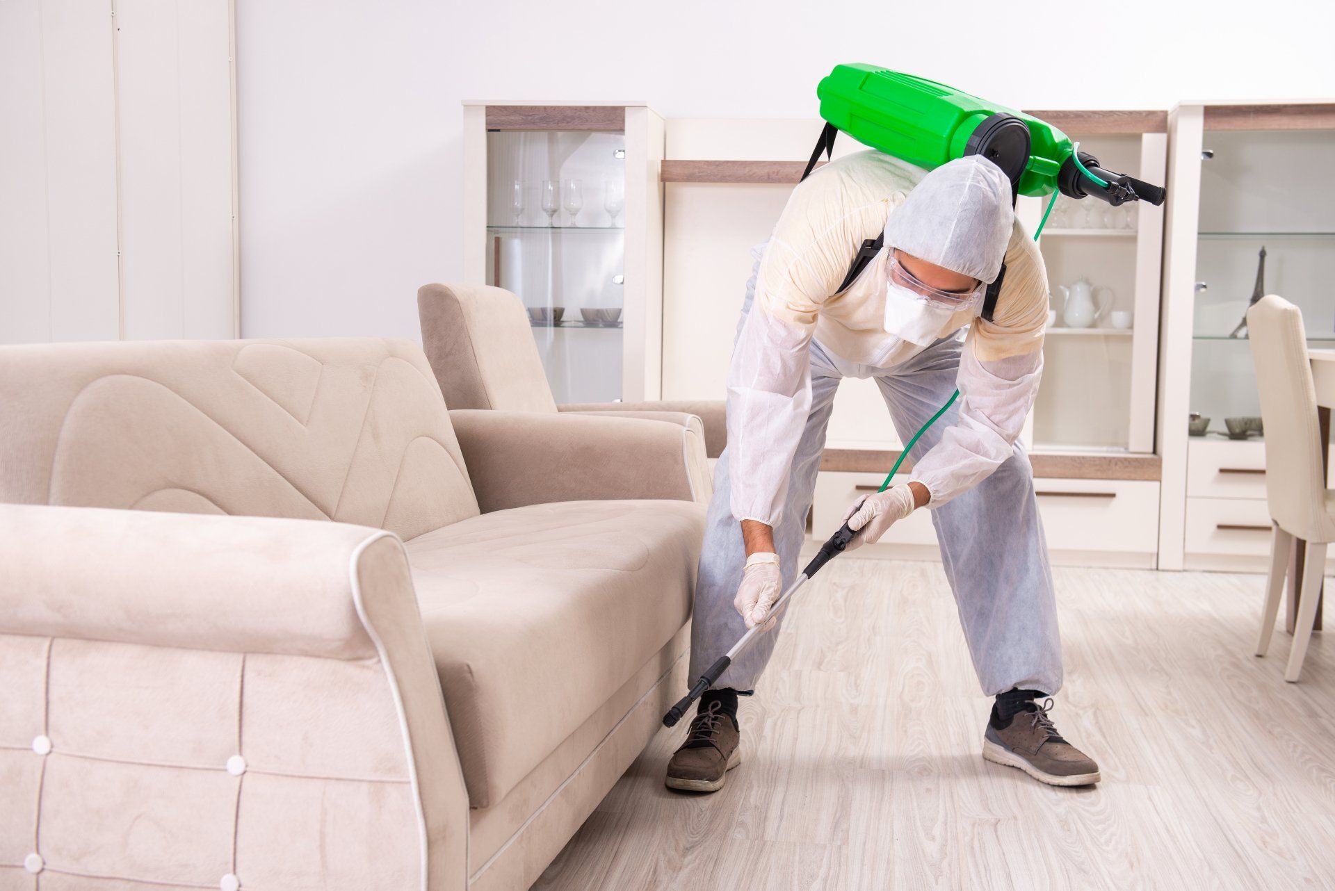 Un technicien en désinsectisation pulvérise un insecticide près d'un canapé beige dans un salon blanc.
