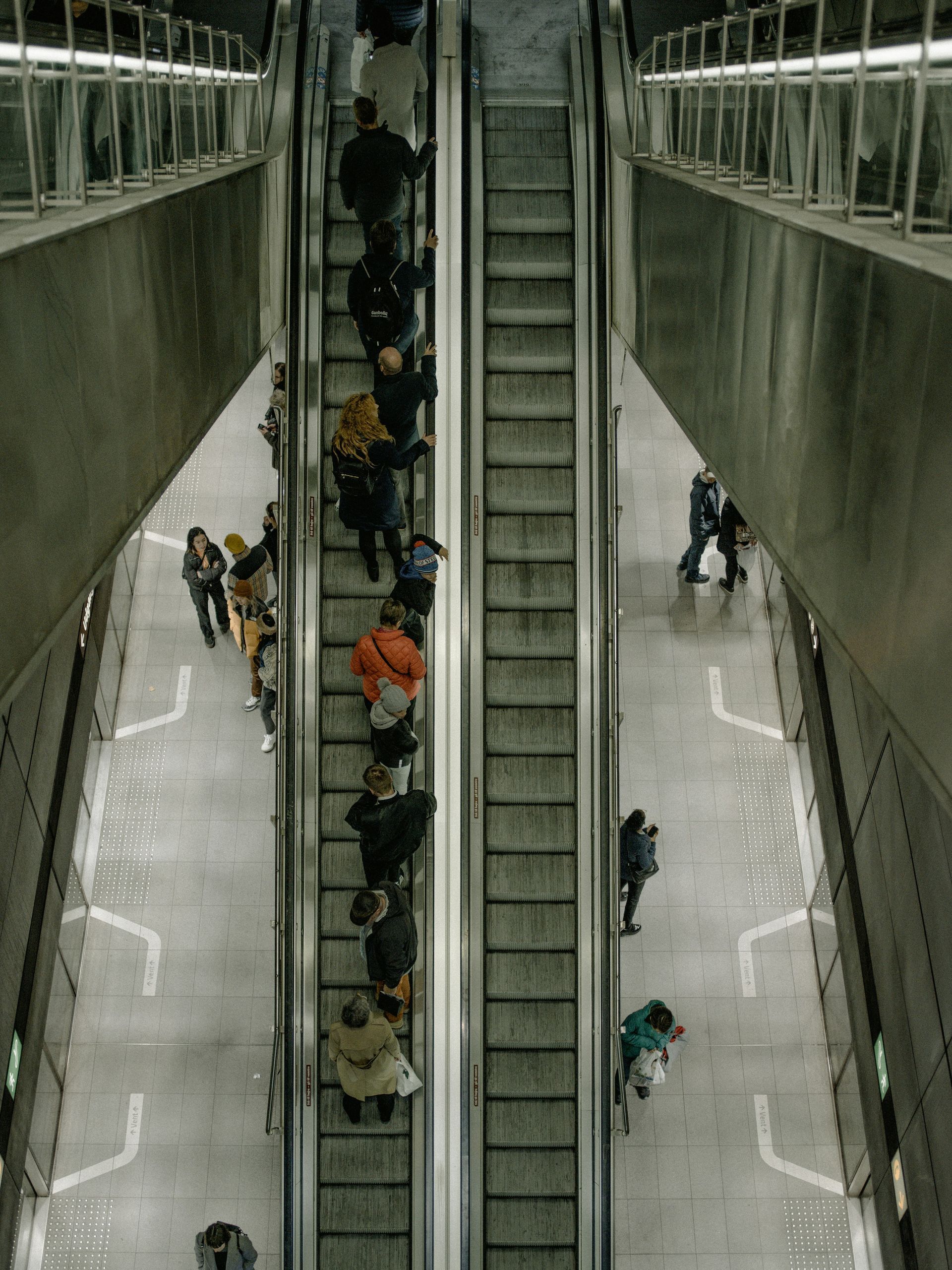 People riding escalators in a building, viewed from above. Concrete floor, metal railings.